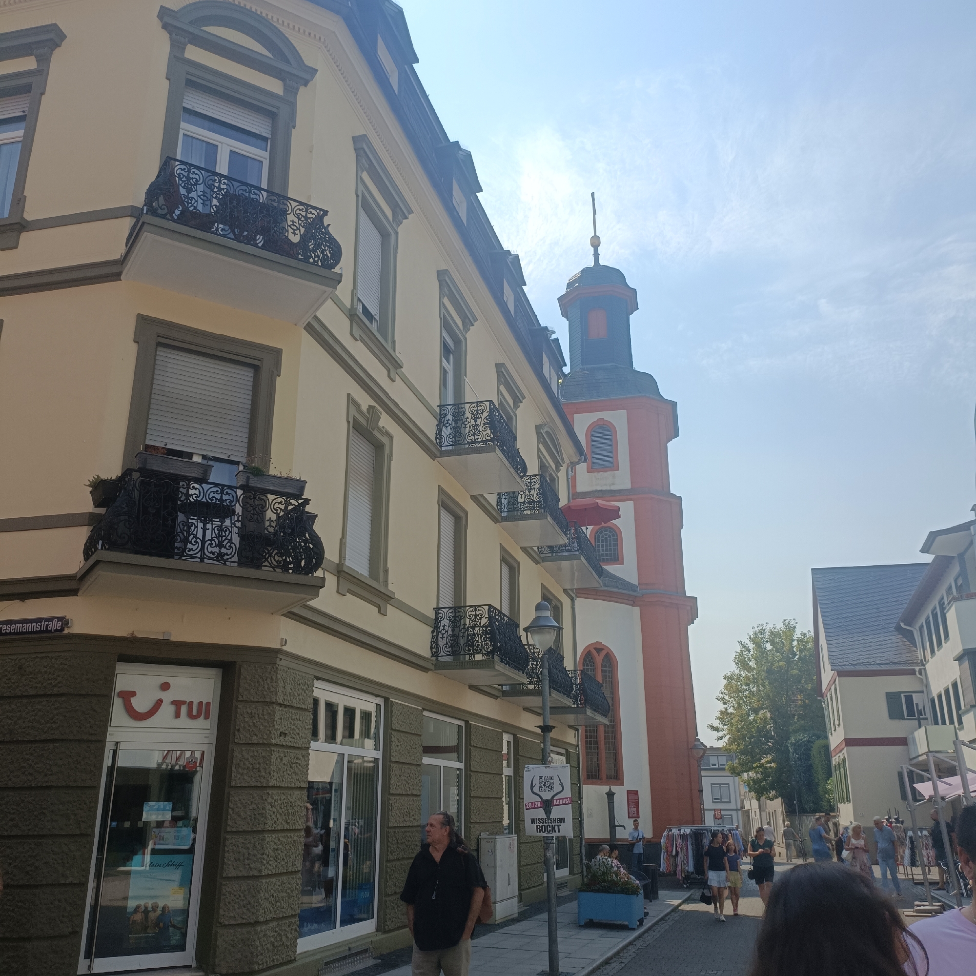 The image depicts a street scene in Bad Nauheim, Germany, with a focus on a building facade and a tower. A pale yellow building with grey trim features multiple windows, some with closed roller shutters, and decorative wrought-iron balconies. A street sign reading "Riesemannstraße" is visible on the left side of the building. A TUI travel agency storefront is located at street level. To the right of the yellow building, a tall, octagonal tower with red and white sections and arched windows rises against a bright, partly cloudy sky. In the foreground, several people are walking along the cobblestone street, with some browsing stalls that appear to be selling clothing. A street lamp with a sign attached is also present. The overall scene suggests a warm, sunny day in a town with historical architecture.