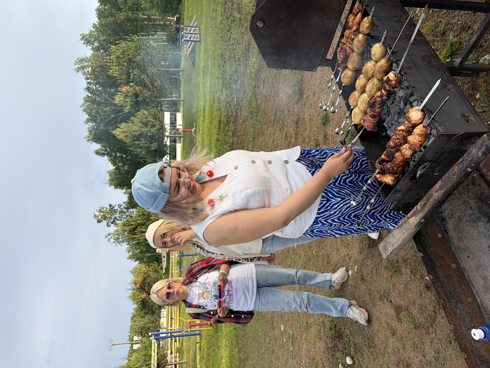 Three women are gathered around a barbecue grill in an outdoor setting. The woman in the foreground, wearing a light-colored sleeveless top and a blue and white patterned skirt, is tending to the grill, which holds skewers of meat and potatoes. To her left, a woman in a white t-shirt and a red and black plaid shirt draped over her shoulders holds a plate with food. She is also wearing light-wash jeans and sunglasses. Behind her, another woman in a cream-colored top and a light-colored hat stands with her hand near her face. The background features a grassy area with trees and what appears to be a playground or park equipment. The sky is overcast. This scene is consistent with a barbecue in a park or recreational area.