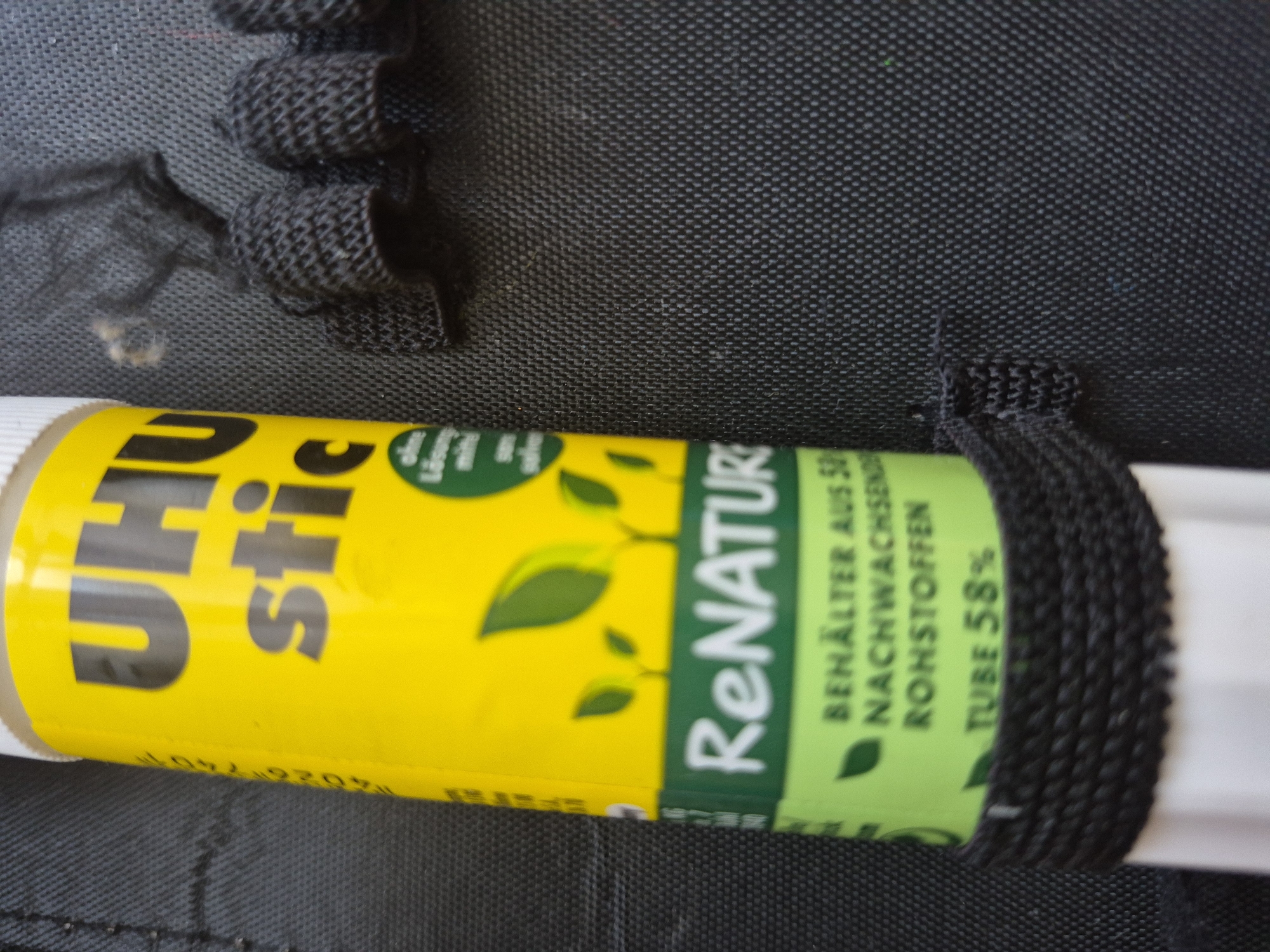 A close-up, slightly angled shot focuses on a yellow and green glue stick, labeled "UHU stick," resting against a textured black surface. The glue stick has a white cap and a black woven band wrapped around its middle. The label features the "UHU stick" logo in large black letters on the yellow portion, with green leaves and sprouts depicted below. The green section of the label includes text that reads "ReNATUR," and further down, "BEHÄLTER AUS 50 NACHWUCHSEN ROHSTOFFEN," and "TUBE 58%." To the right of the glue stick, several black, woven cylindrical objects are visible, partially obscuring the background. The black surface appears to be a fabric with a distinct woven pattern. No people or city elements are present in the image.