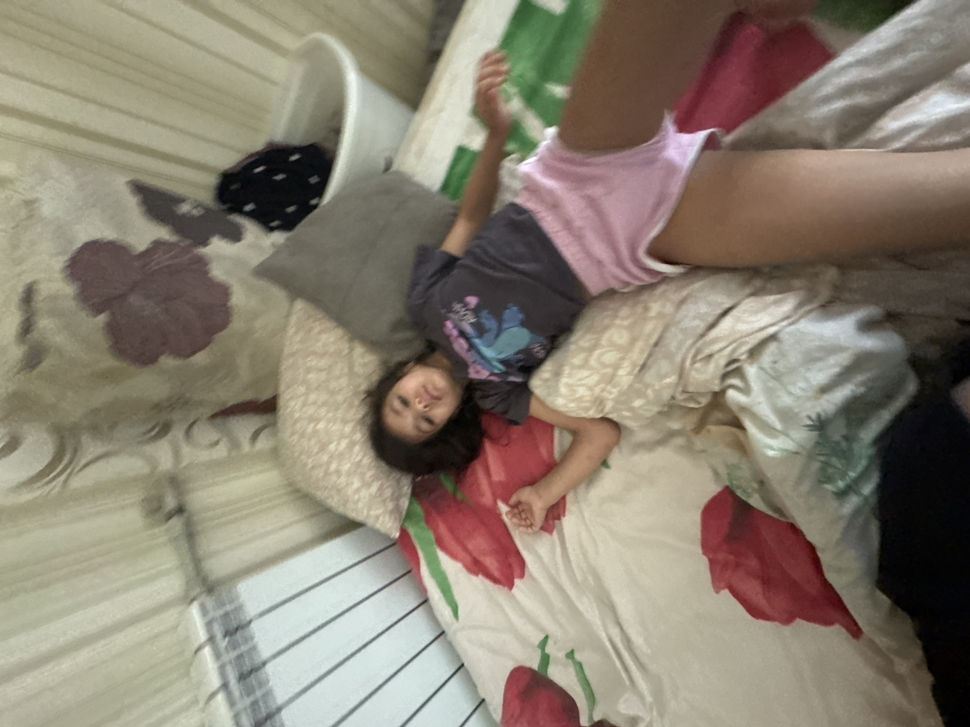 A young girl with dark hair is lying on a bed with floral patterned sheets. She is wearing a grey t-shirt with a blue character on it and pink shorts. The room has striped curtains and a radiator. There is a pillow behind her and to her left. In the background, there is a white hamper with some dark fabric inside. The overall lighting suggests it is daytime. The image appears to have been taken indoors in a residential setting, possibly a bedroom.