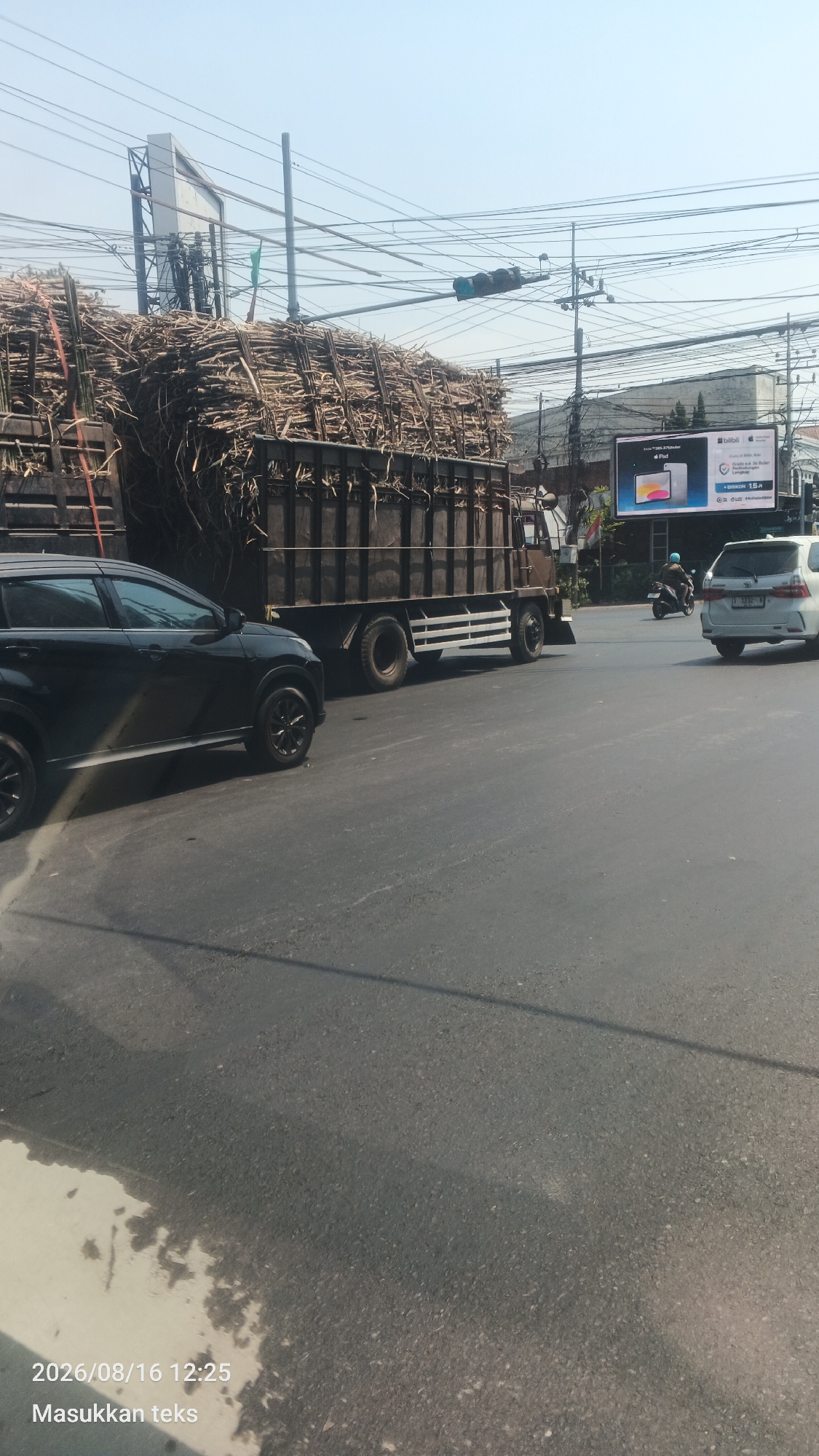 A large truck loaded with sugarcane stalks is parked on the side of a road, with a black car parked in front of it. The truck is dark brown with a white stripe along the bottom. The sugarcane stalks are dry and brown. A billboard is visible in the background, showing an advertisement for an iPad. A traffic light is visible on a pole above the road. A white car and a motorcycle are also visible on the road. The sky is clear and blue. The scene appears to be in a city, possibly in Malang, Indonesia, given the timestamp and text overlay in Indonesian.