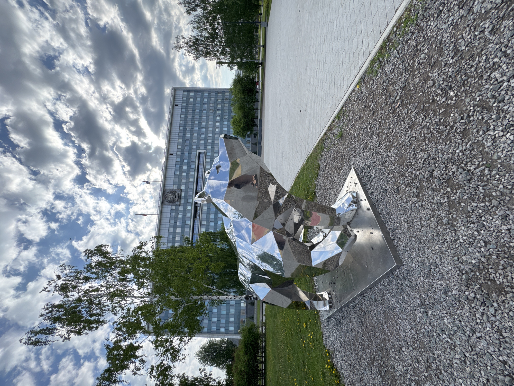 The image depicts a modern, geometric-shaped, shiny metal sculpture of a bear in a park setting. The sculpture sits on a metal base, which is placed on a gravel-covered ground. A large, multi-story building is visible in the background, with a sign featuring a circular emblem and a flag on top. Green trees are visible on either side of the building. The sky is partially cloudy. This scene is located in Perm, Russia.