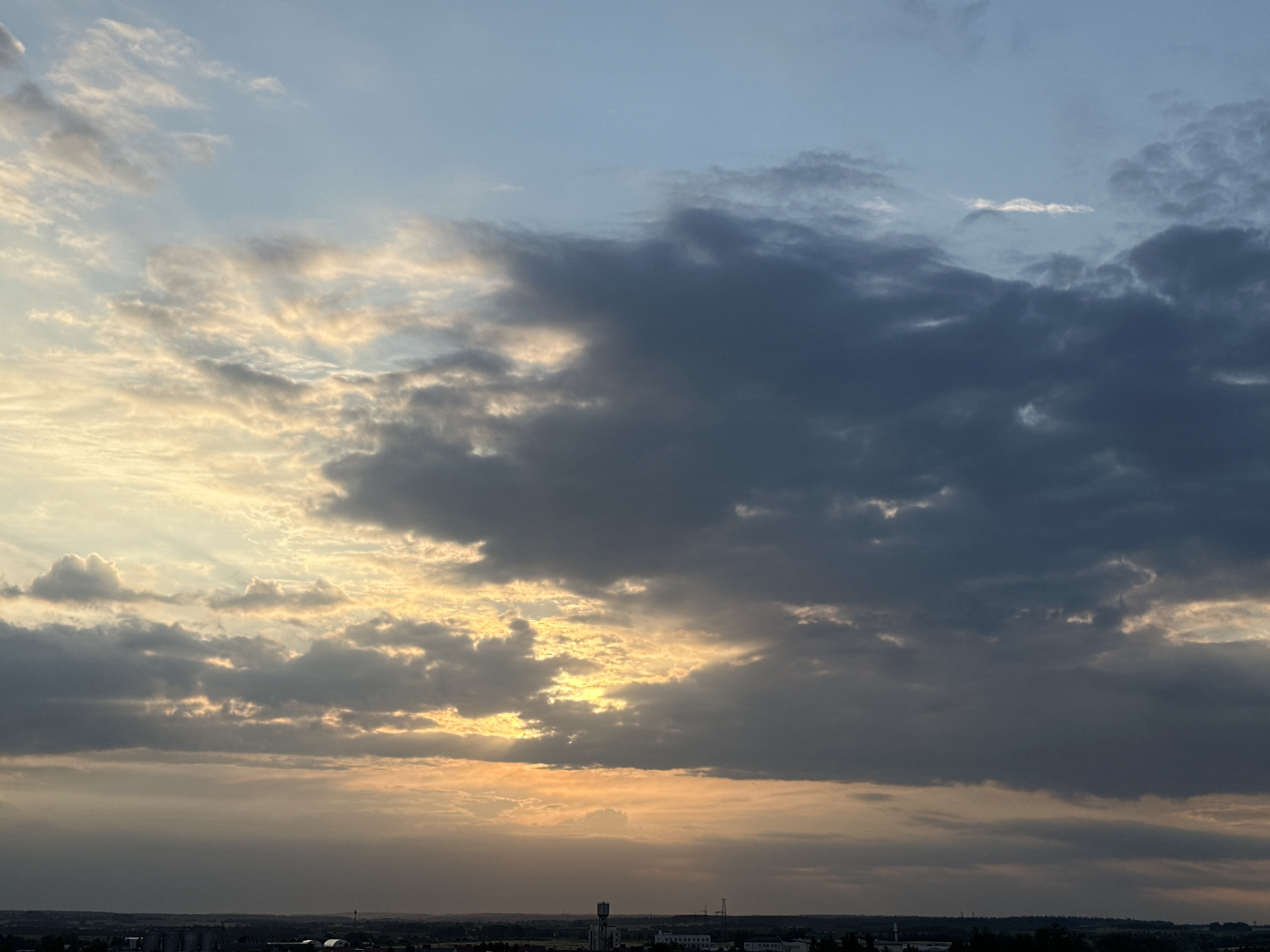The image depicts a wide shot of a sky at sunset over the city of Shchomyslitsky Sel'sky Soviet, Belarus. The sky is filled with a mix of dark and light clouds, with the setting sun casting a warm, golden glow through breaks in the clouds. The lower portion of the image shows a dark silhouette of the city's skyline, with a few buildings and structures visible. The overall atmosphere is dramatic and serene.