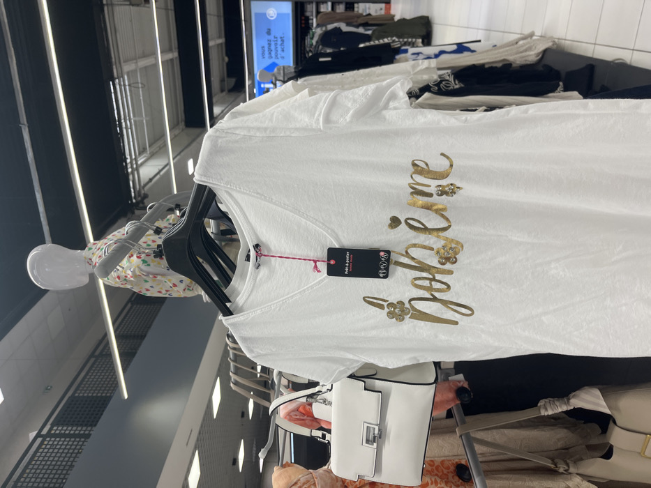The image shows a white t-shirt with a gold "bohème" inscription on a hanger inside a clothing store. The store is located in Saint-Brice-Courcelles, France, at coordinates 49.283 latitude and 4 longitude. There is a white purse hanging next to the t-shirt. In the background, a mannequin is wearing a multi-colored shirt. Several other garments hang on hangers in the background. The clothing store appears to be well-lit and well-organized. The image does not include any people. A sign in French is visible in the background advertising a potential prize for a purchase. The t-shirt has a small pink tag hanging from it.
