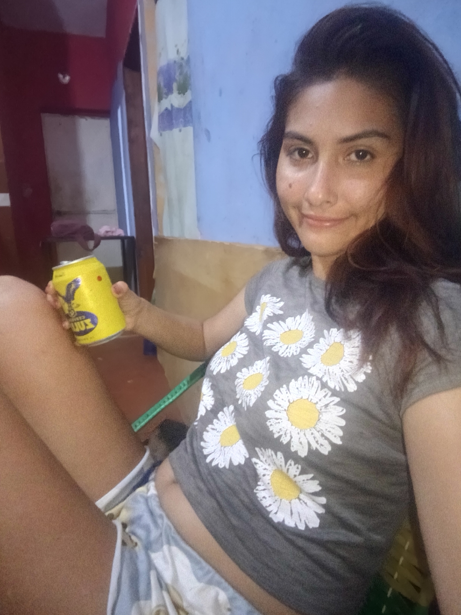 A woman is sitting indoors, holding a yellow can with a blue and black logo. She is wearing a gray shirt with white and yellow daisy prints and patterned shorts. In the background, there is a doorway leading to another room with a table and various items, and a blue wall with a curtain hanging on it. The location is Juan Griego, Venezuela.