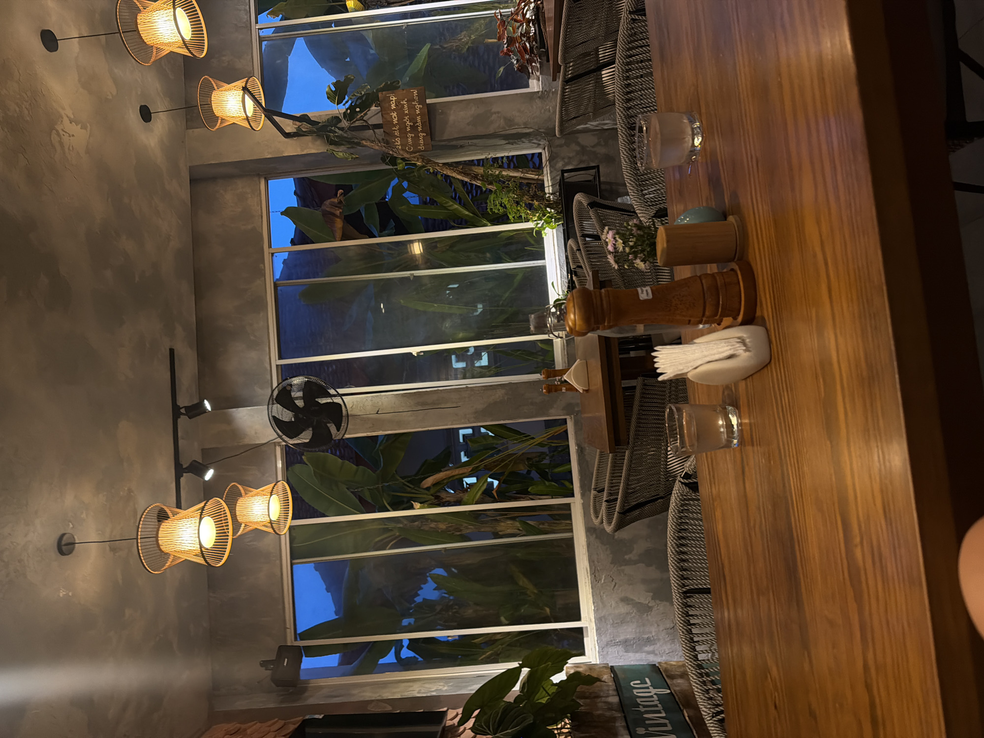Here's a detailed description of the image:
**Location:** The scene appears to be an indoor space, likely a restaurant or cafe, in Nha Trang Ward, Vietnam. This is suggested by the presence of Vietnamese text on signage visible through the windows.
**Environment:** The space has a modern, possibly rustic, design. The walls are a textured gray, and large windows look out onto lush greenery and blue sky, creating a bright and airy atmosphere. The interior is well-lit by decorative light fixtures with a woven, lantern-like design. A ceiling fan is also visible.
**Objects:**
* **Table:** A long wooden table dominates the right side of the image. It's set with what seems to be a minimalist arrangement of glassware, salt and pepper shakers, napkins, and small floral decorations.
* **Chairs:** Woven chairs, probably made of rattan or a similar material, are lined up along the table.
* **Signage:** There are signs with Vietnamese text visible through the windows. One sign is attached to some greenery. Another says "vintage" on it.
* **Plants:** There are many green plants, both inside and outside the windows, adding a natural, tropical element to the scene.
**Overall:** The image captures a stylish and inviting indoor space with a connection to the outdoor environment through the large windows and abundance of plants. The presence of dining settings indicates a place for eating and socializing.