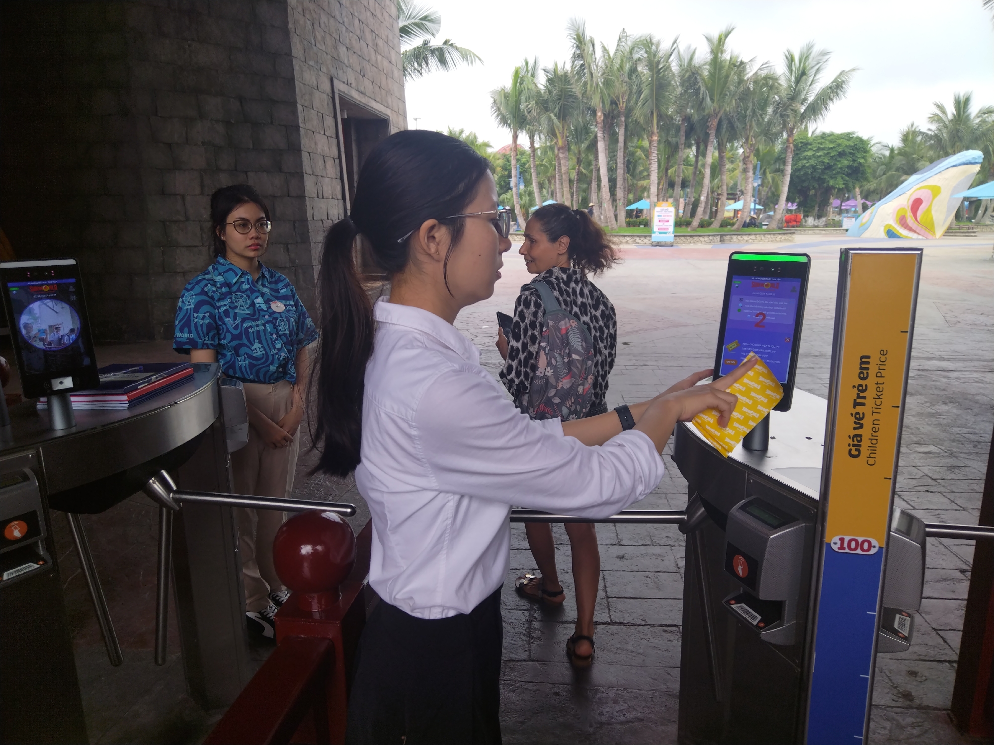 The image shows the entrance to a water park in an unknown city. A young woman in a white shirt and black skirt is inserting a yellow ticket into a ticket scanner to pass through a turnstile. Behind her, a woman with a ponytail and a spotted shirt is standing. To the left of the turnstile, a staff member in a patterned blue shirt stands near a table with a ticket scanner. The area is paved with stone, and in the background, there are palm trees and a large, colorful wave sculpture.