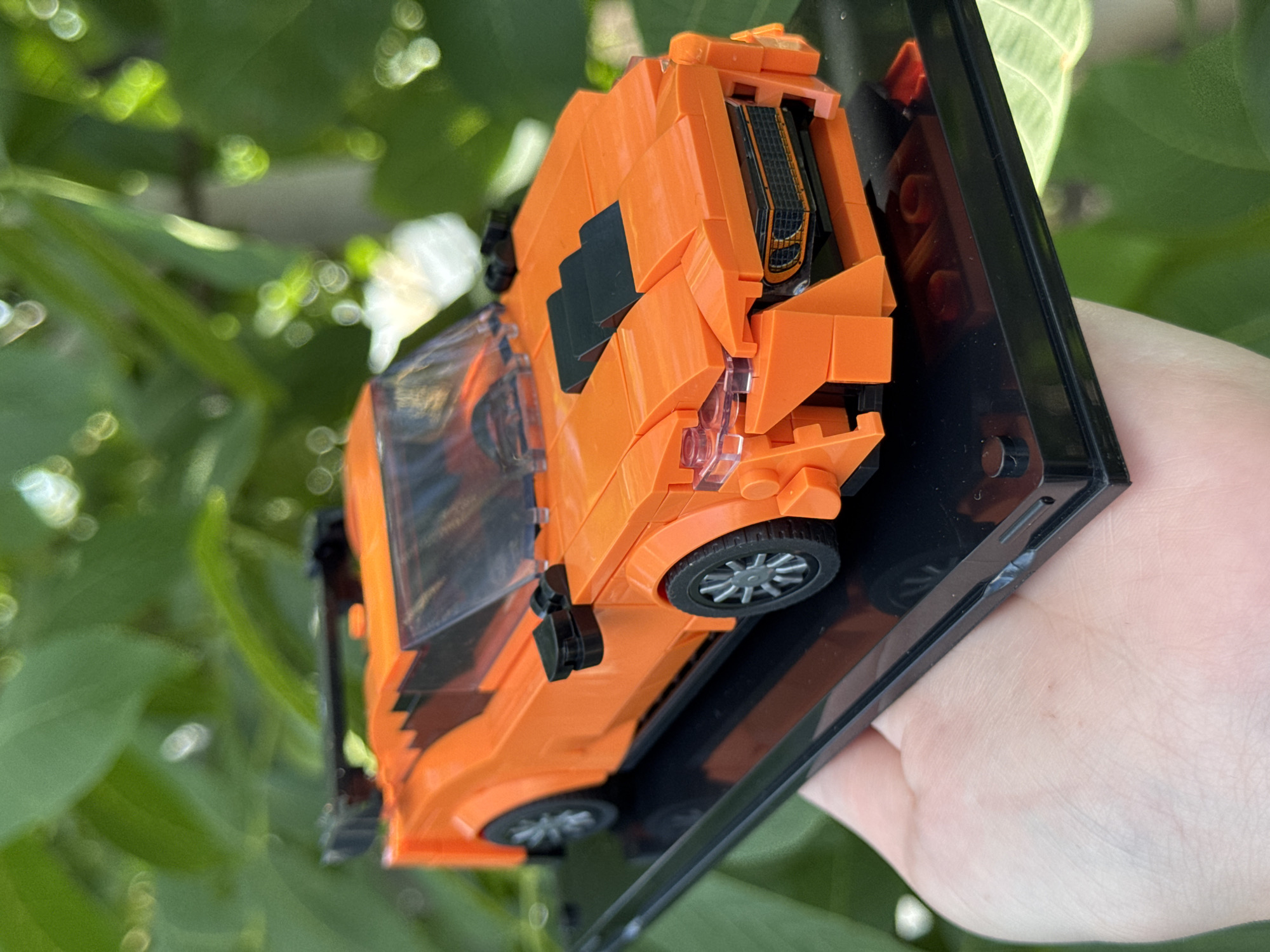 This media file shows a close-up, angled shot of an orange Lego car model, presented on a black display base. The car is intricately constructed from various orange Lego bricks, featuring clear windows, black accents, and realistic silver-rimmed wheels. The detailed construction suggests a focus on replicating a sports car or a similar vehicle. The Lego car is held by a person's hand, partially visible on the right side of the frame, with their thumb and fingers gently supporting the display base. The background is blurred, revealing lush green foliage and bright, out-of-focus highlights, indicating an outdoor setting, possibly a garden or park. The natural lighting and soft bokeh effect suggest a sunny day. There is no visible text in the image.
