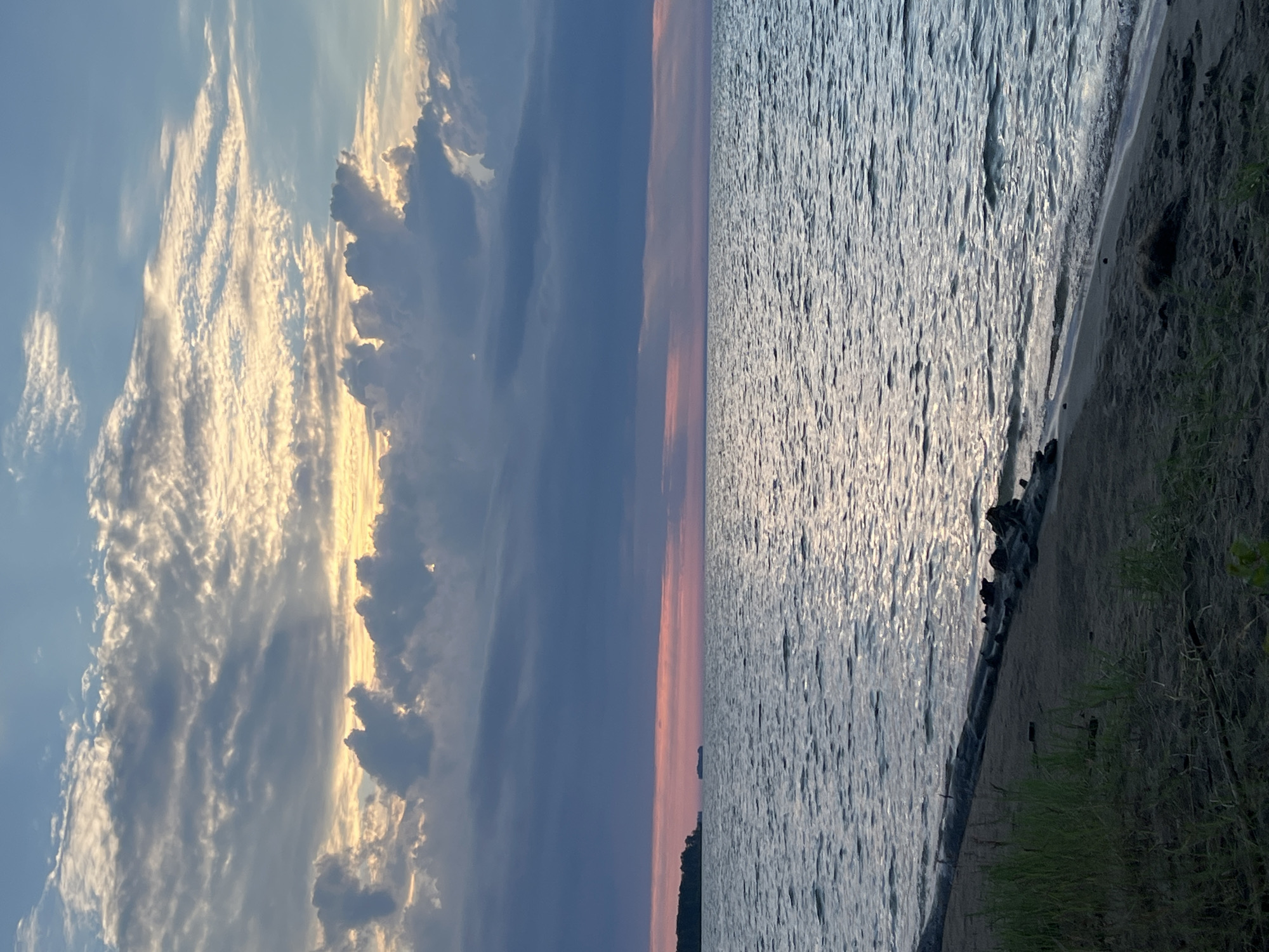 The image depicts a serene sunset over a body of water. The sky is filled with dramatic clouds, ranging from dark blue and purple hues to lighter, golden-tinged formations illuminated by the setting sun. A thin band of pink and orange hues is visible along the horizon, indicating the sun's descent. The water is calm, with gentle ripples reflecting the sky's colors and the light of the setting sun. The foreground shows a sandy shore with some driftwood and sparse green vegetation. In the distance, a dark, tree-covered landmass is visible on the left, with a small structure or object out in the water. The overall scene suggests a peaceful and natural environment. The location is identified as the city of Borovoe, Russia.