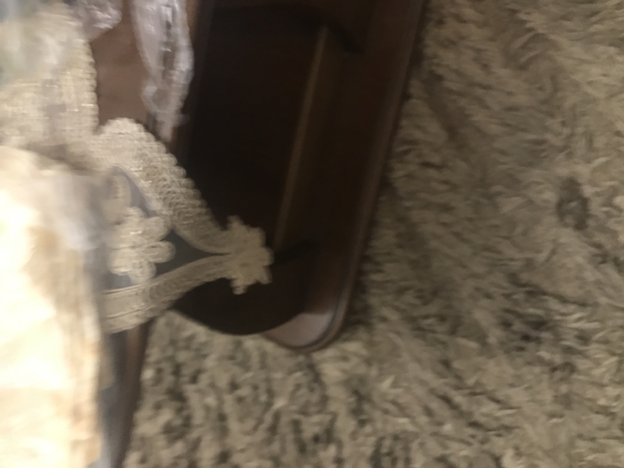 The image shows a portion of a white lace-trimmed object, possibly fabric, resting on or near a dark brown wooden frame. The background appears to be a light-colored, textured rug or carpet. The location is not identifiable from the given image.
