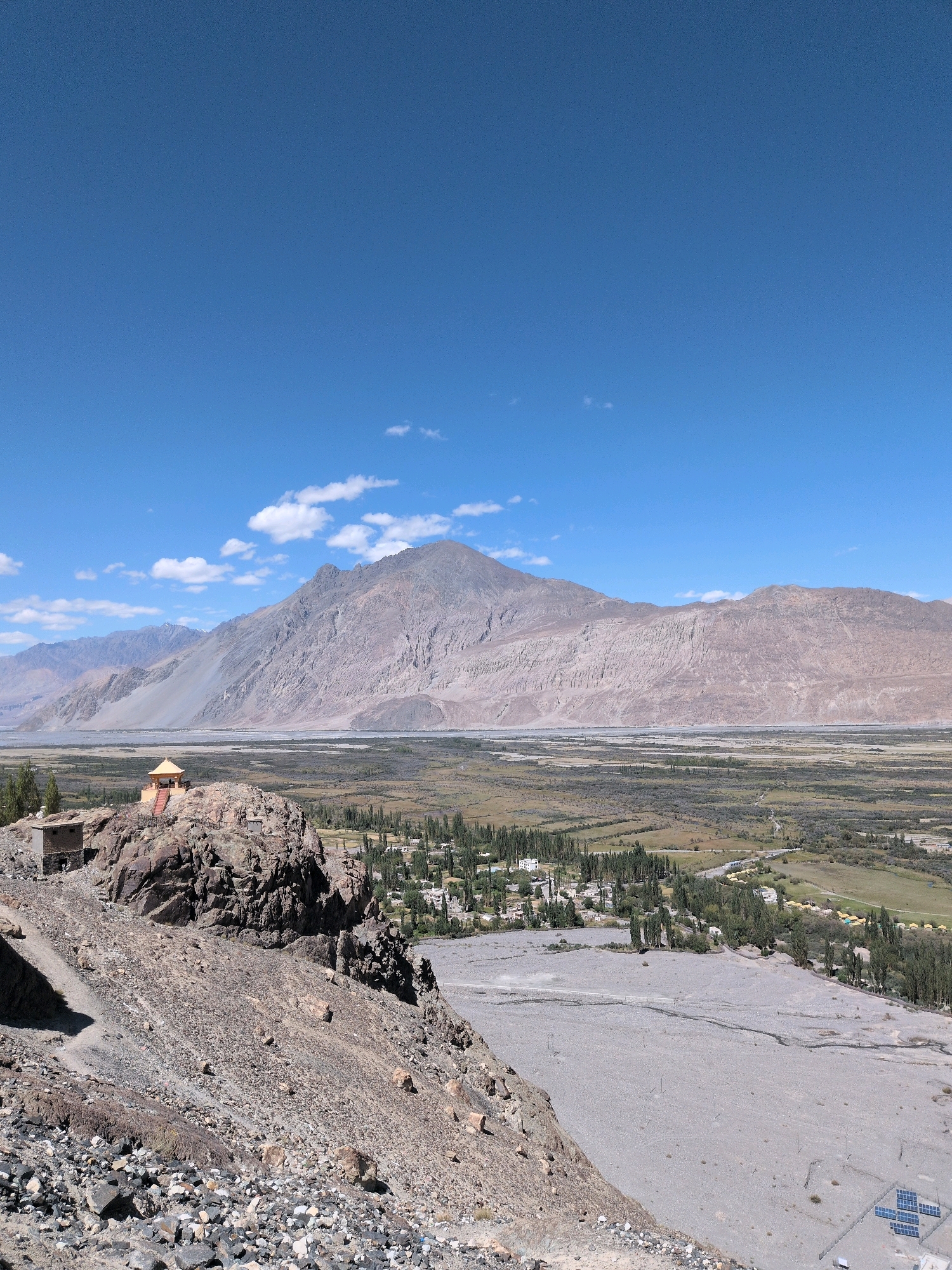 An aerial, wide-angle shot captures the expansive, rugged terrain surrounding Diskit, India. The sky is a vibrant, clear blue with a few wispy white clouds. In the foreground, a rocky, arid hillside slopes downwards, dotted with loose stones and gravel. A narrow, winding path is visible on the left. Perched atop a prominent rocky outcrop is a small, yellow-roofed structure with red stairs leading up to it. Next to this structure, on the same rock formation, is a small, square, stone building.
Further into the midground, a lush green valley stretches out, punctuated by rows of tall, slender trees. A village with white-roofed buildings is nestled within this green expanse, and a grey, dry riverbed cuts through the landscape. In the background, imposing, barren mountains rise majestically, their textured slopes displaying shades of brown and grey. The scene is devoid of any human presence.