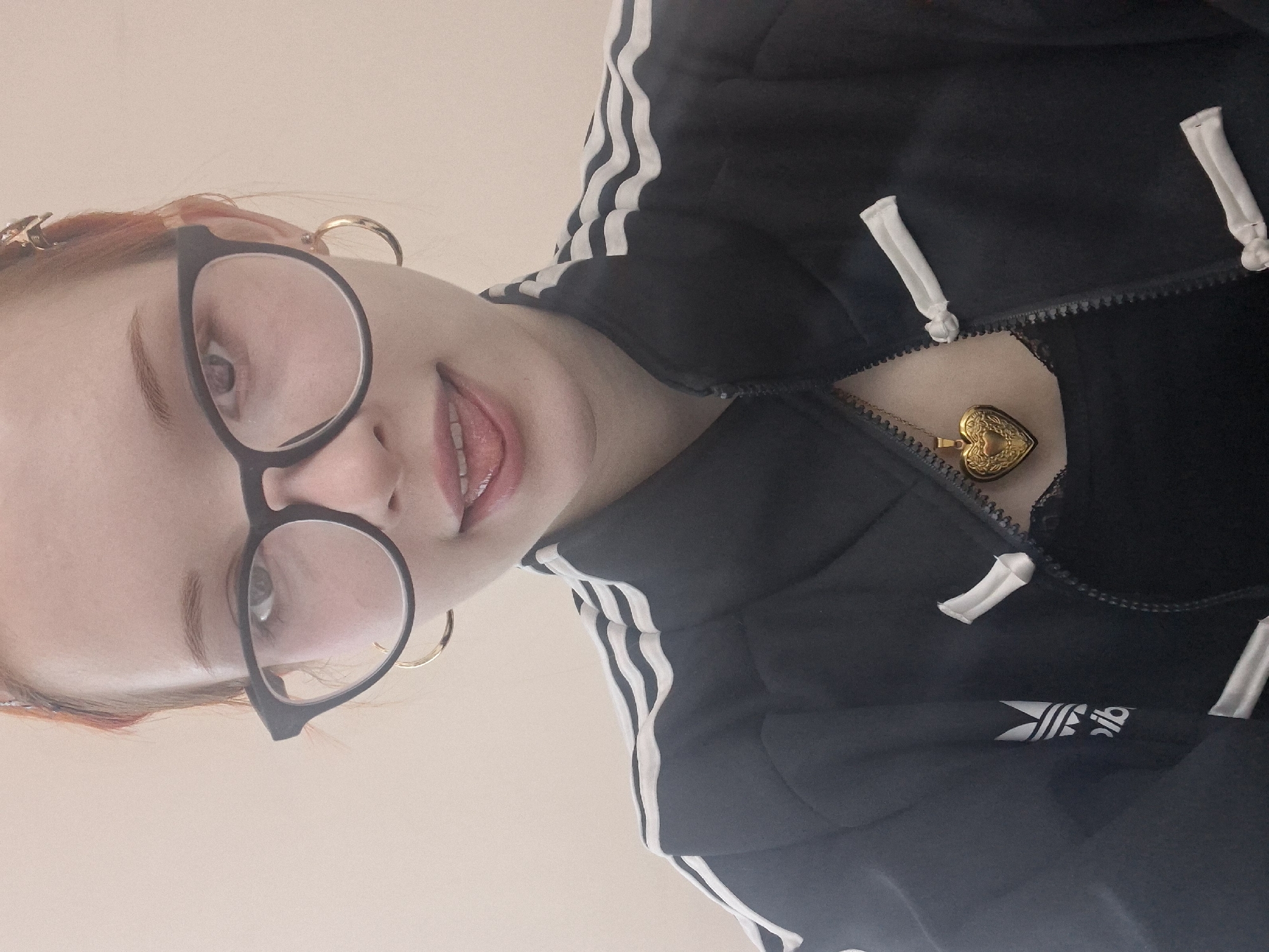 A woman with fair skin, round glasses, and red hair poses for a selfie. She is wearing gold hoop earrings and a black Adidas jacket with white stripes. A gold heart-shaped locket hangs from her neck. The background is a light beige color. There is no indication of the location being in Chaikovsky, Russia.