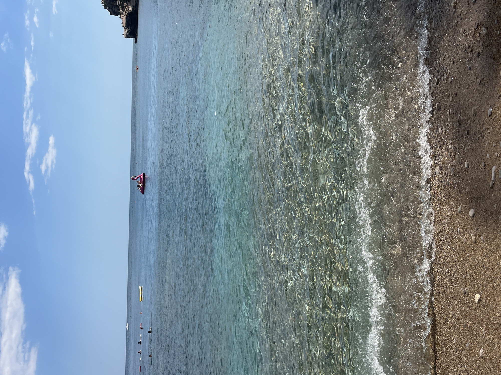 The image shows a daytime scene at a beach in Budva, Montenegro. The water is clear, transitioning from deeper blue further out to a crystalline turquoise near the sandy shore. Gentle waves are washing onto the beach, which is composed of small pebbles and sand. In the distance, there's a rocky headland. Further out, a pink inflatable flamingo with people on it is visible on the water's surface. There are other people swimming further out. The sky is a clear blue with a few scattered white clouds.