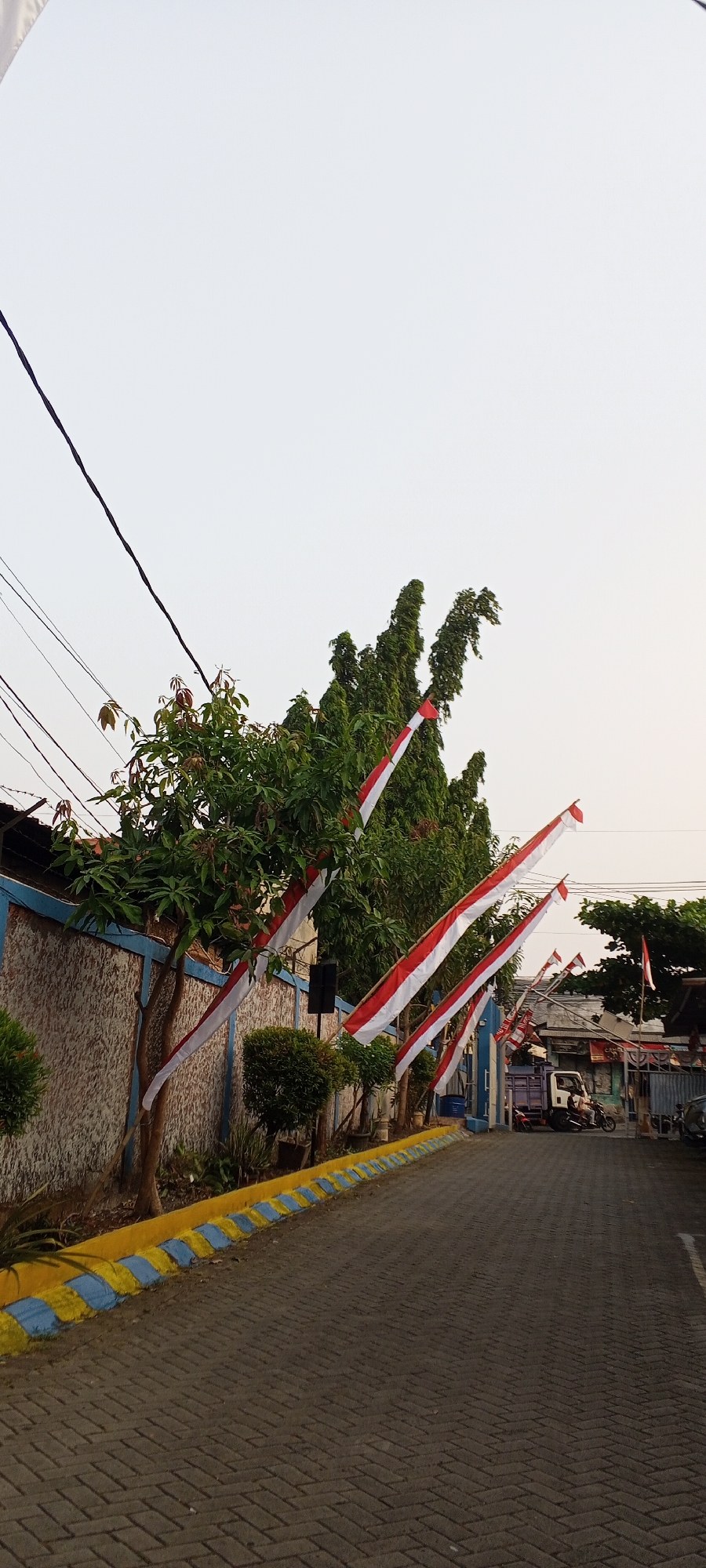 The image shows a street in Depok, Indonesia, decorated with Indonesian flags. The flags are red and white, and they are hung on poles along the side of the road. The street is paved with interlocking bricks, and there is a wall on the left side of the road. There are also trees and bushes along the side of the road. In the background, there are buildings and vehicles, including a truck and a motorcycle. The sky is overcast. The flags suggest that it may be a national holiday or a patriotic event.