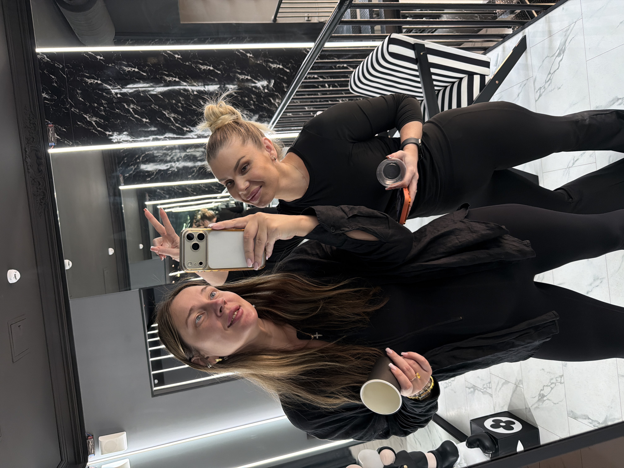 Two women are taking a selfie in what appears to be a modern interior space, possibly a boutique or a stylish lounge area. The woman on the left, with long brown hair, is holding a dark paper cup in her left hand and her right hand is extended towards the camera. She is wearing a black long-sleeved shirt and black leggings. The woman on the right, with blonde hair in a bun, is holding a smartphone in her left hand and a small circular object in her right hand. She is wearing a black long-sleeved top and black leggings. Both women are smiling and looking at the phone's screen. In the background, there is a black and white striped chair, a staircase with a metal railing, and reflective surfaces that suggest a mirror or polished walls. The environment features modern lighting, including vertical light strips. There are no visible elements that indicate a specific location in Shcherbinka, Russia.