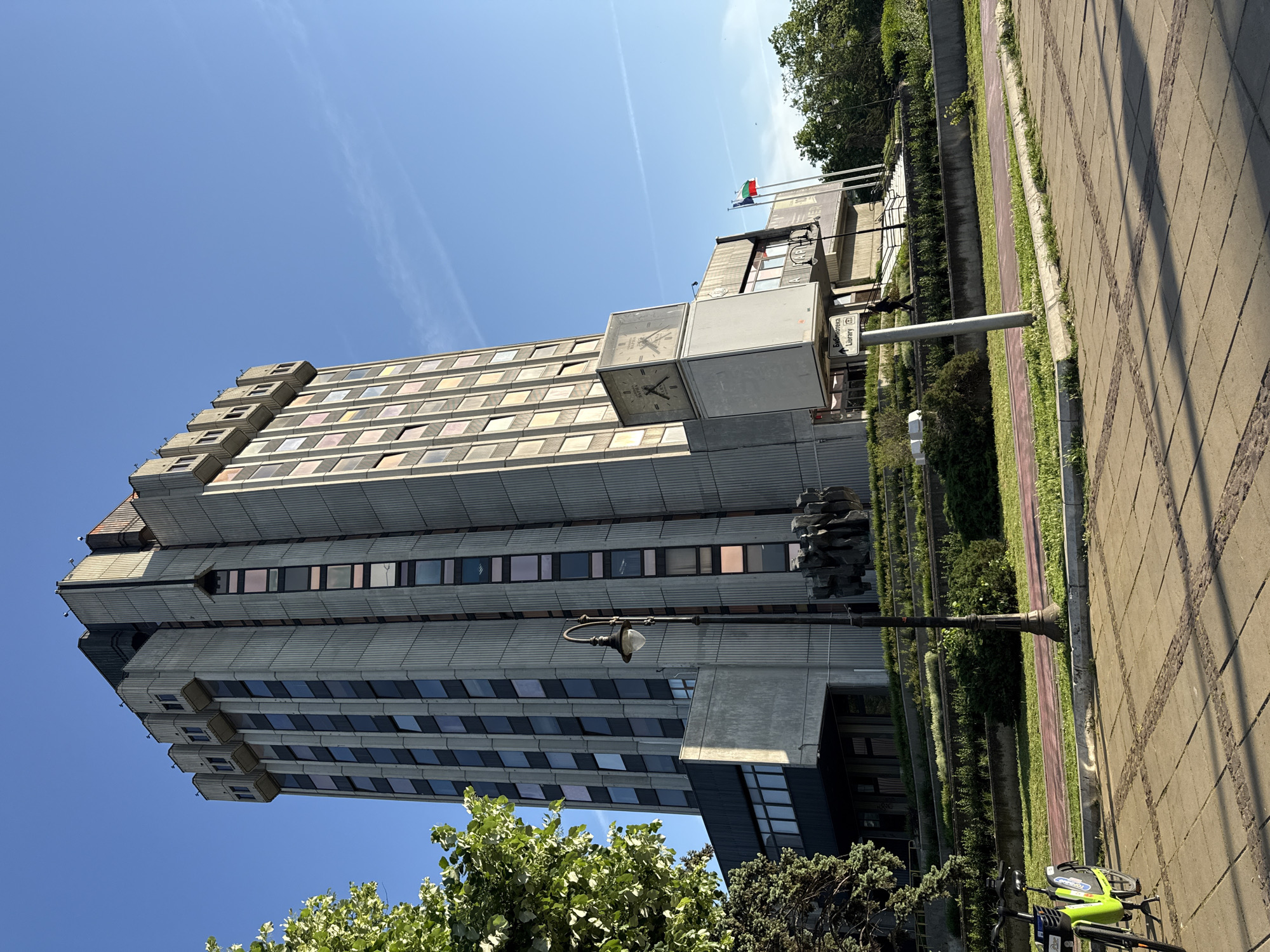 Here is a forensic description of the image:
The image shows a tall, imposing building with a modernist, Brutalist architectural style. The building appears to be a government or public services building, possibly a library, given the signage. The building's facade is characterized by horizontal bands of windows, and concrete. A clock tower is affixed to the side of the building.
The background includes blue sky and some trees. The foreground shows a pedestrian walkway, a green bike, and some greenery.
Based on details like the flag and signage, the location is in the city of Sofia, Bulgaria. The building appears to be the National Library of Bulgaria "St. Cyril and St. Methodius".