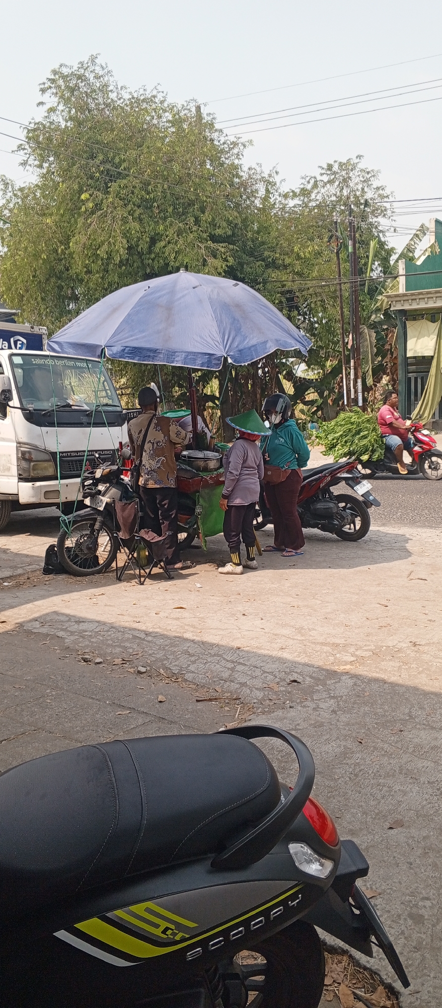 In Maospati, Indonesia, a group of people are gathered under a large blue umbrella by a roadside stall. A white truck is parked to the left of the stall. A motorcycle is parked in front of the stall, and another motorcycle is seen in the background with a person on it. The stall itself appears to be selling food items. The ground is paved, and trees are visible in the background. The sky is clear and bright.