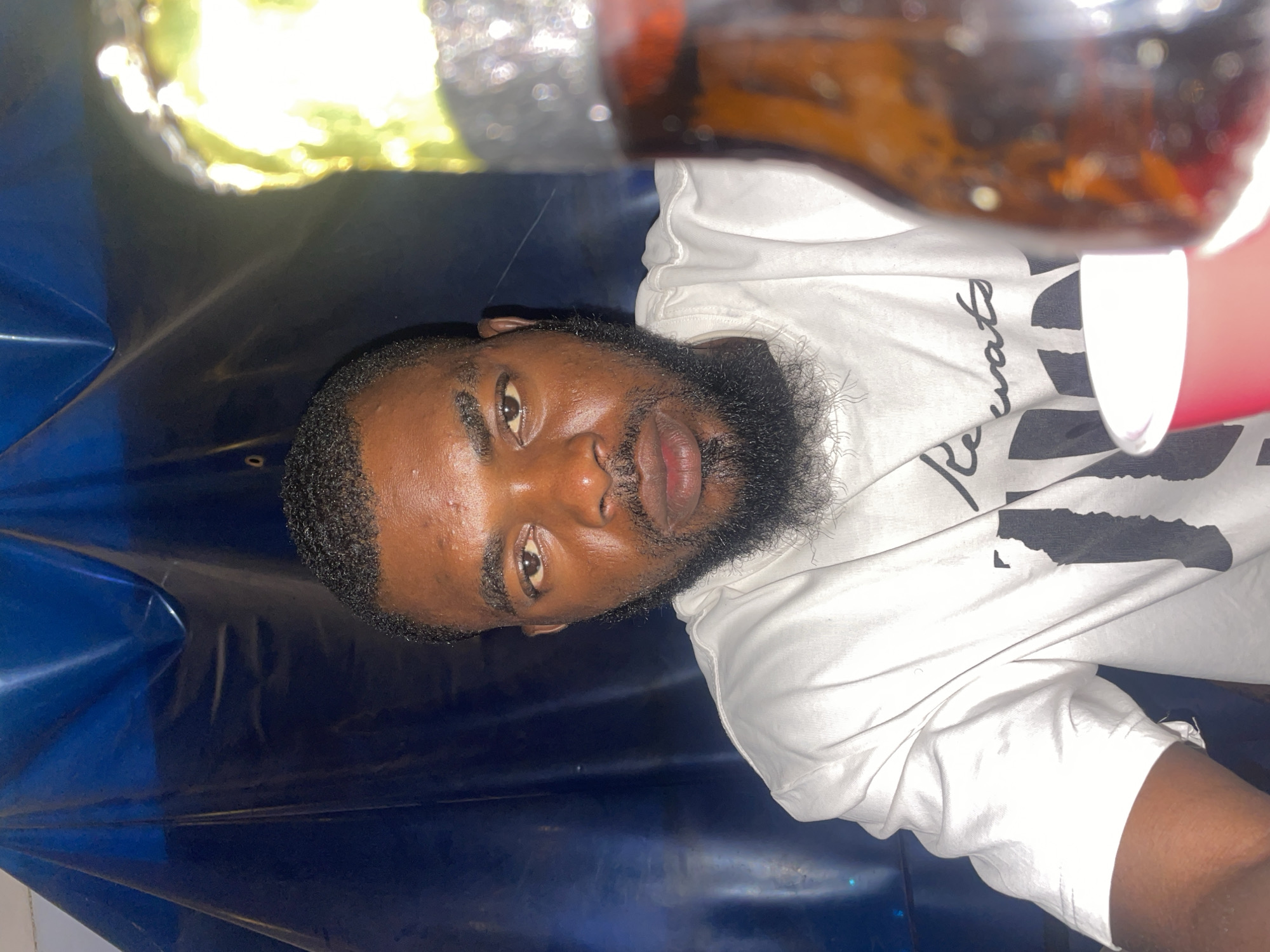 A selfie of a man with dark skin, a short haircut, and a beard is taken from a low angle. He is wearing a white t-shirt with black lettering. In the upper center of the frame, a brown bottle with a gold foil wrapper is visible, partly obscuring the background. The background appears to be a shiny blue and black surface. The location and city of Jos, Nigeria, are not definitively discernible from the provided visual data.