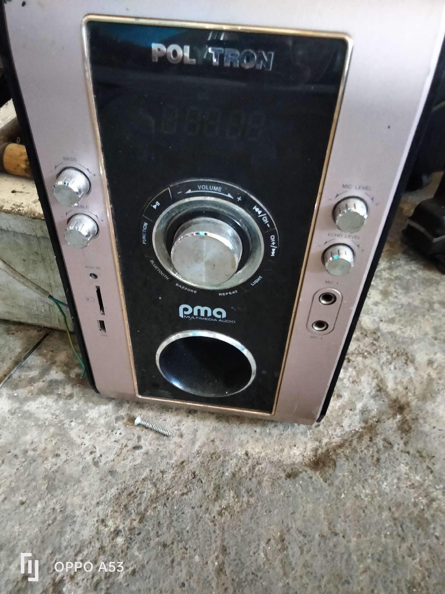 A portable multimedia audio device, identified by the brand "POLYTRON" and model "pma" on its front panel, is situated on a concrete surface. The device features various controls, including knobs labeled "BASS," "TREBLE," "VOLUME," "MIC LEVEL," and "ECHO LEVEL." It also has input ports for "AUX IN" and "USB," as well as two microphone jacks labeled "MIC 1" and "MIC 2." The surrounding environment appears to be outdoors, with debris and an electrical wire visible on the ground. The image was captured in Denpasar, Indonesia, as indicated by the "OPPO A53" watermark, a common smartphone brand in that region. The lighting suggests it is daytime.
