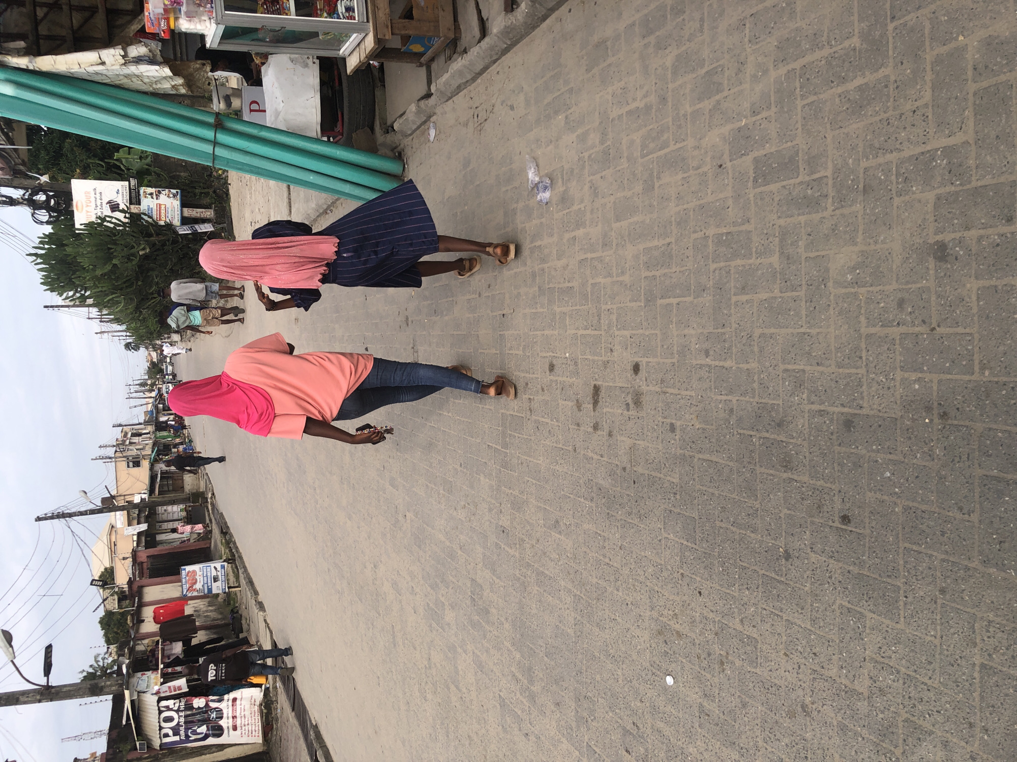 The image depicts a street scene in Ado, Nigeria. Two young women, one in a pink headscarf and coral top, the other in a pink headscarf and a navy blue striped dress, walk away from the camera down a cobblestone street. The woman on the right is holding a beaded item. Other people and structures line the street, including shops, advertisements, and streetlights. In the background, a man in a black shirt and blue jeans walks away from the camera. A row of tall, green pipes stands vertically to the right of the two women. The sky is overcast.
