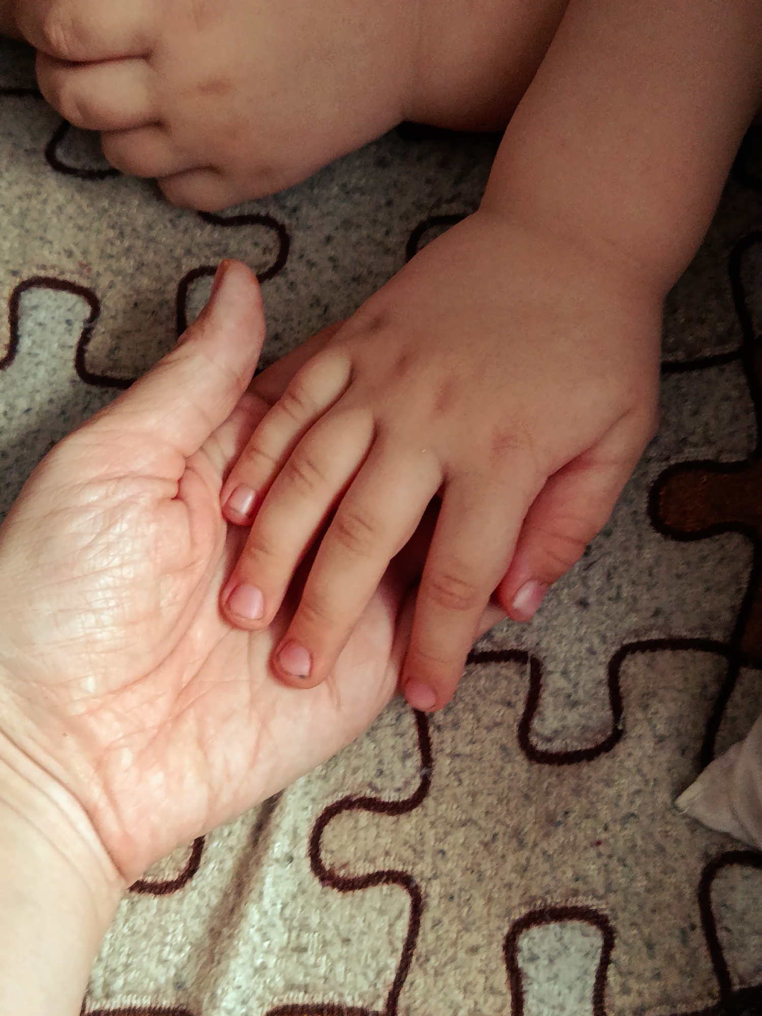 This vertical close-up photograph captures a tender interaction between an adult and a child. In the foreground, an adult's open hand, characterized by visible lines and wrinkles, gently holds a smaller, smoother child's hand. The child's fingers rest softly on the adult's palm, revealing short, light-colored fingernails. Portions of the child's plump arm or leg are also visible in the upper and right parts of the frame.
The hands are positioned over a textured textile, likely a blanket or rug, which features a distinct brown puzzle-piece pattern against a light, mottled background of grey and beige tones. The warm, muted color palette suggests an indoor setting, possibly a home. The gesture conveys a sense of connection, care, and protection between the two individuals. There are no discernible details to indicate the time of day or specific weather conditions. The setting for this image is Фоки, Russia. No visible text is present in the media.