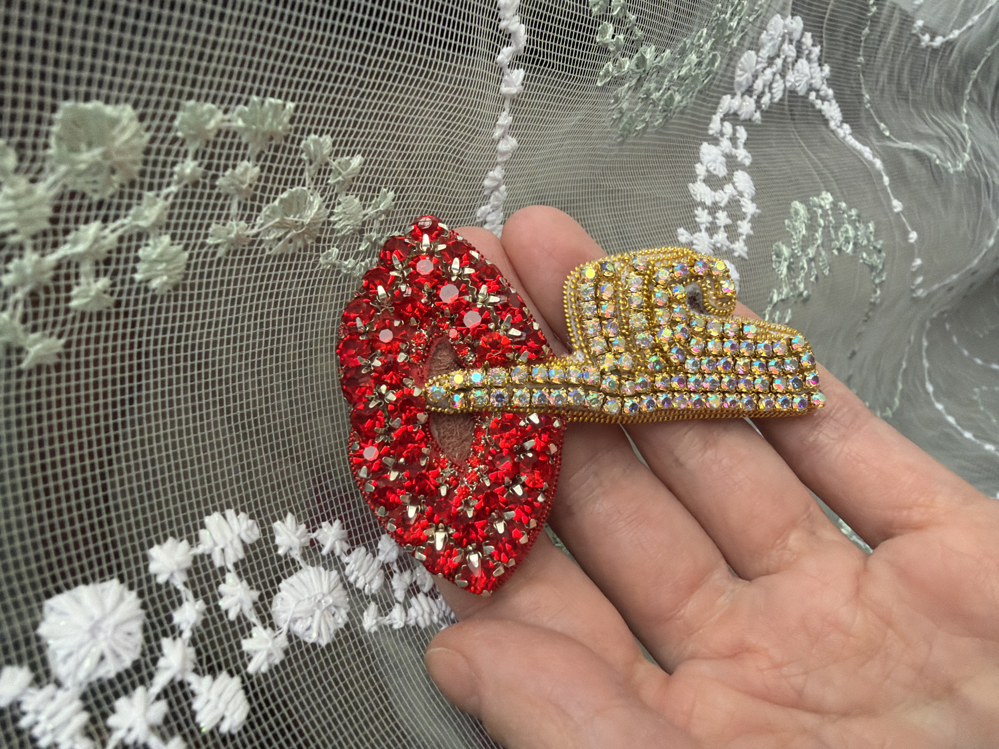 A person's hand holds a red and gold jeweled brooch over a sheer fabric background with white and green floral embroidery. The brooch consists of a red heart shape and a rectangular gold section covered in small jewels. The person's hand is in focus.