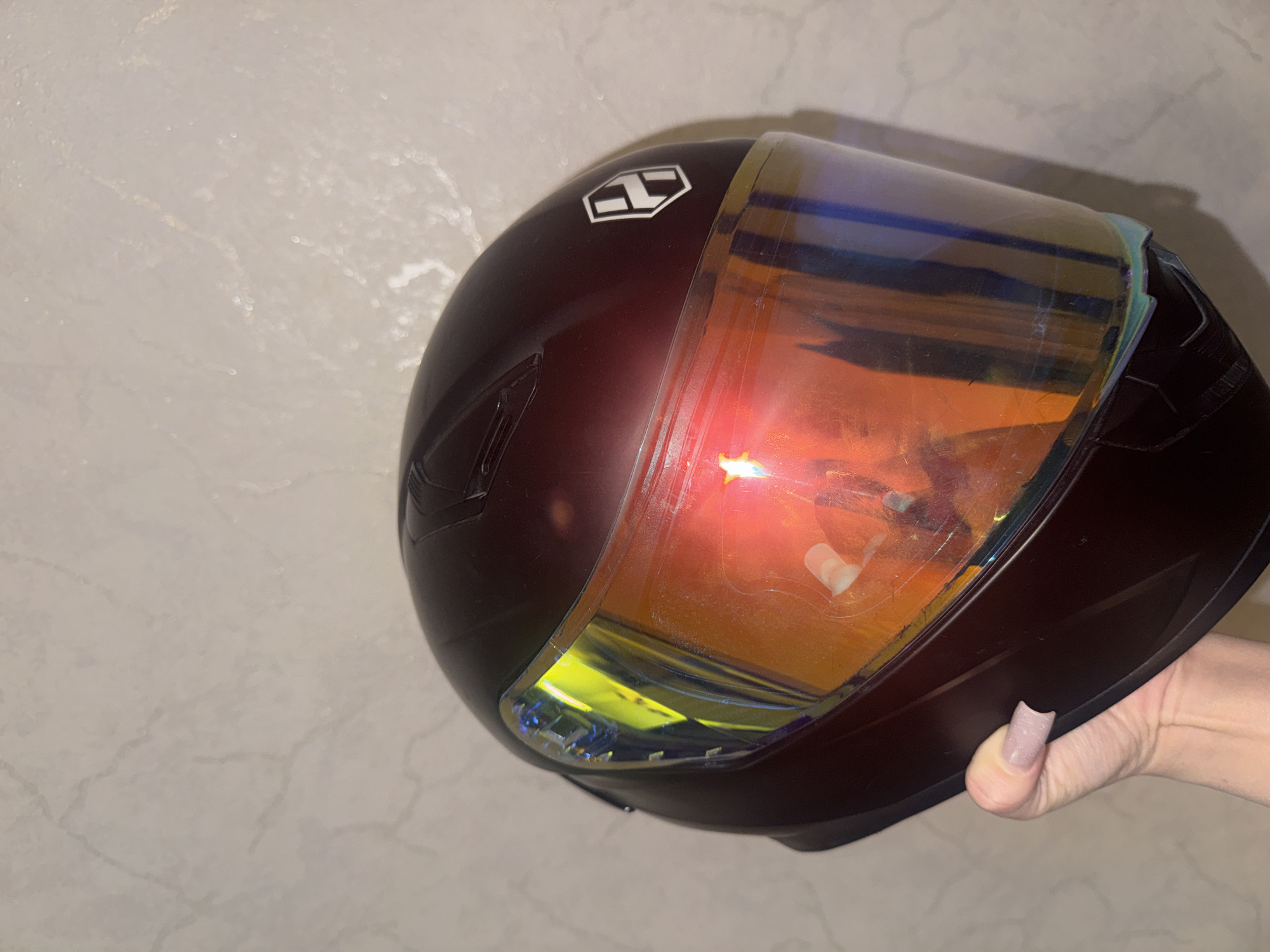 A person's hand is holding a dark red motorcycle helmet. The helmet has a reflective visor that displays hues of yellow, orange, and blue. A white logo with a stylized 'H' is visible on the side of the helmet. The background is a neutral-colored, textured wall, resembling marble. There are no discernible landmarks to indicate a specific location within Bishkek City, Kyrgyzstan.