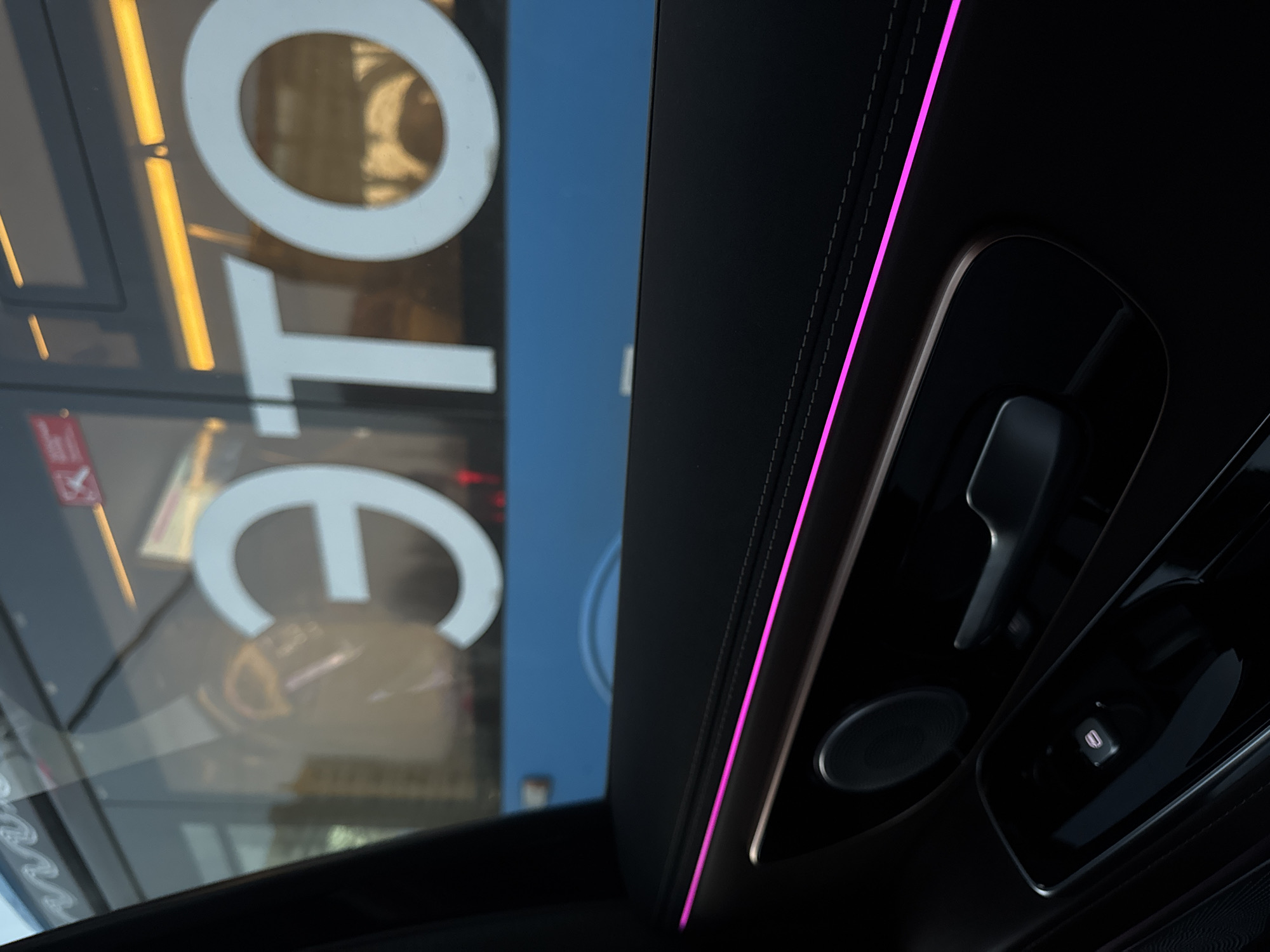 The image shows the interior of a car, specifically the sunroof area and the passenger-side controls. A pink LED strip highlights the edge of the car's ceiling. The passenger-side controls include a reading light and a handle. The car's sunroof is open, revealing a view of the exterior. The reflection on the sunroof glass shows large, white Cyrillic letters, "СТО," which is likely part of a sign. There's also a red sign visible through the sunroof. Given the Cyrillic letters, it's likely that the location is in Russia, possibly in Moscow. The reflection suggests a commercial establishment nearby, possibly a car service or repair shop ("СТО" is a common abbreviation for "Service Station" in Russian).