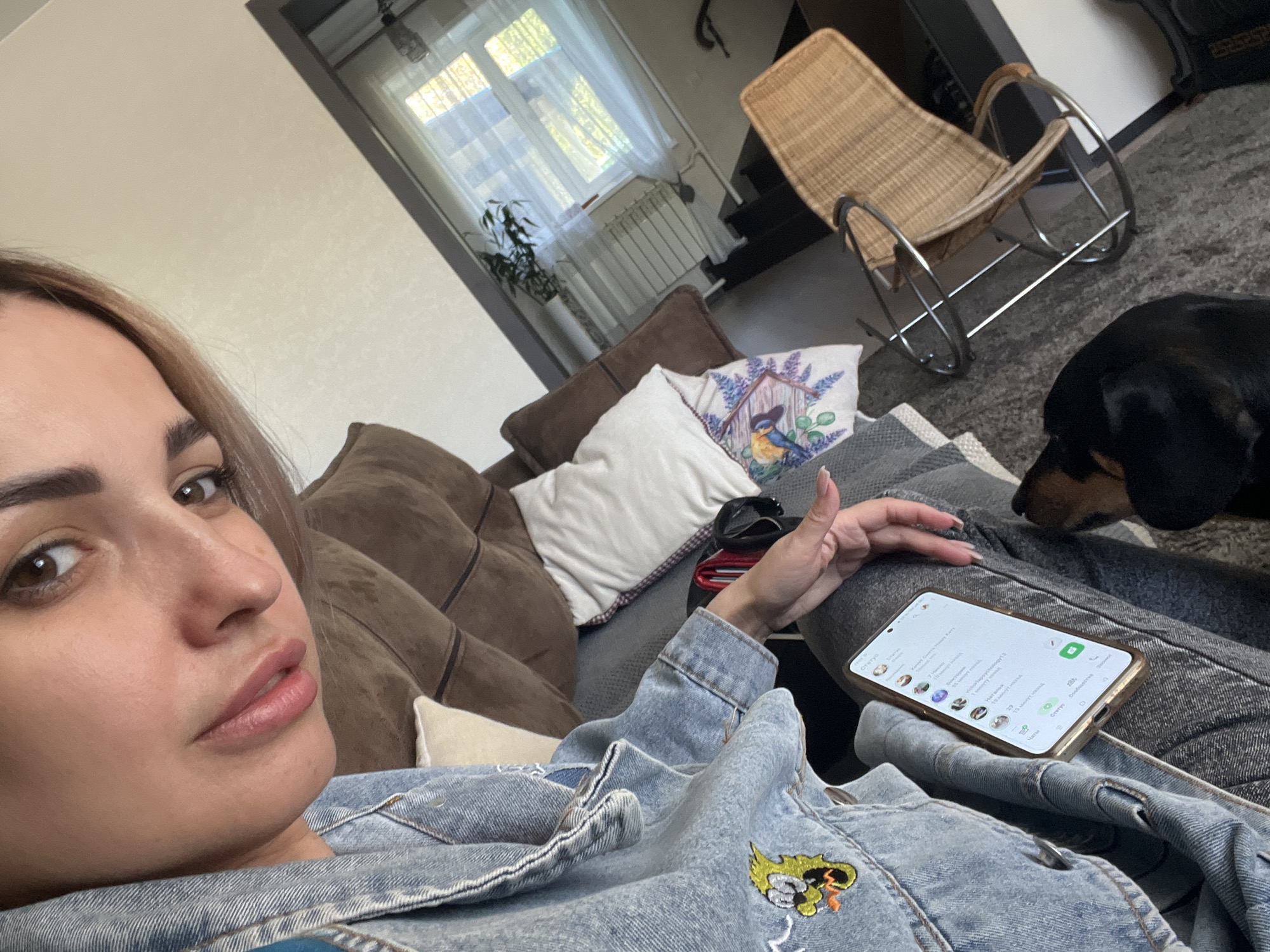 A woman lies on a brown sofa, looking at the camera while holding a phone on her lap. A dachshund dog is near her, sniffing. Pillows are on the sofa, and a wicker rocking chair is visible in the background, near a window with white curtains. The location appears to be inside a residential building.