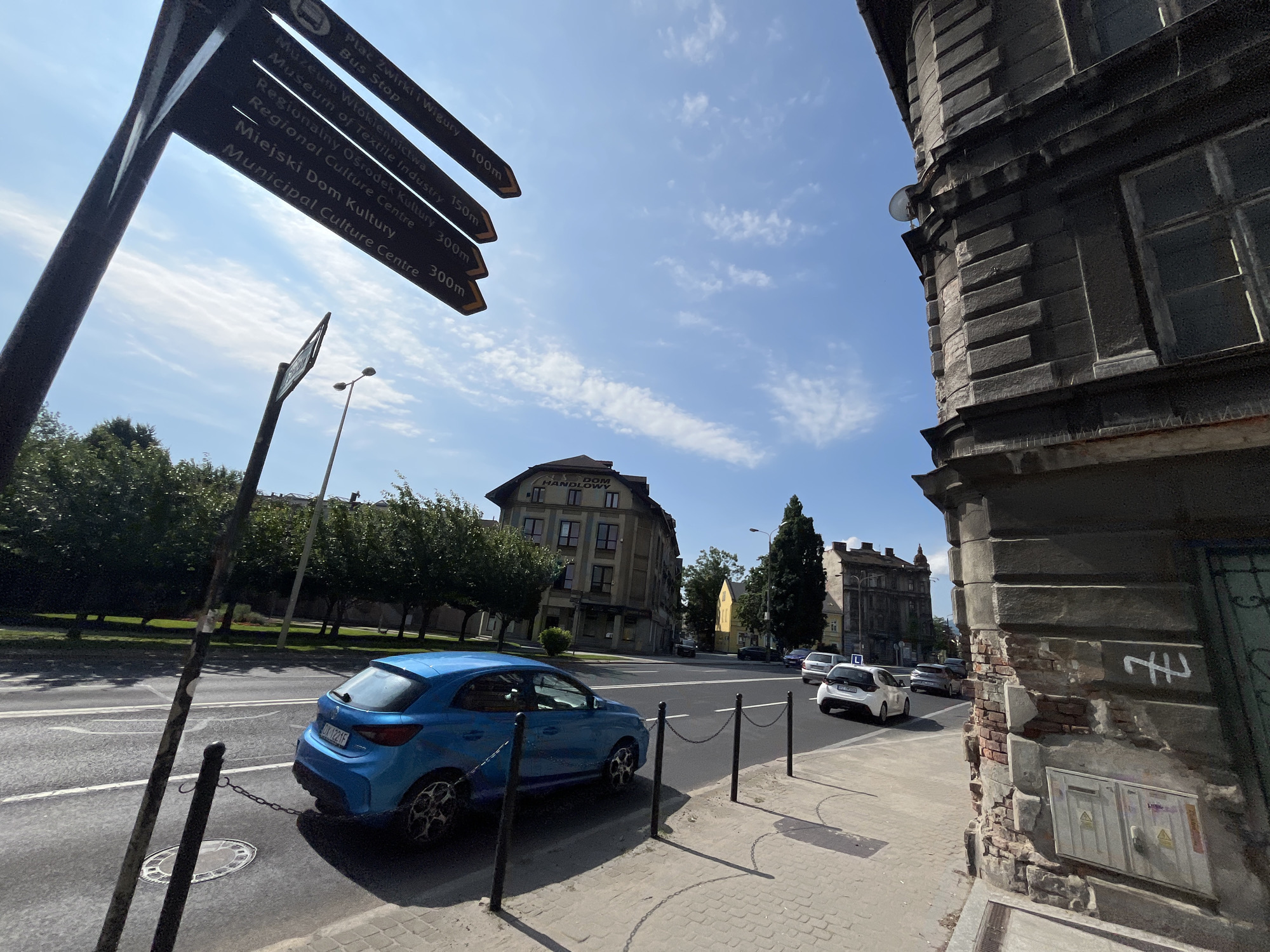 The image depicts a street scene in a city, featuring a blue car parked near a sidewalk and a street sign pointing towards various locations. The street sign is black with white lettering and arrows, indicating distances to places like "Plac Zwrinki i Wigury," "Bus Stop," "Museum of Textile Industry," "Regionalny Ośrodek Kultury," and "Miejski Dom Kultury."
The blue car is a hatchback, facing away from the camera. It is parked next to a black metal chain fence that separates the sidewalk from the road. The sidewalk is paved with cobblestones.
In the background, there are several buildings and trees. One building in the mid-ground appears to be a multi-story structure with a sign that reads "DOM HANDLOWY." Across the street, another building with elaborate architectural details is visible, partially covered by the edge of the frame. The sky is blue with scattered white clouds. The overall impression is a sunny day in an urban environment.