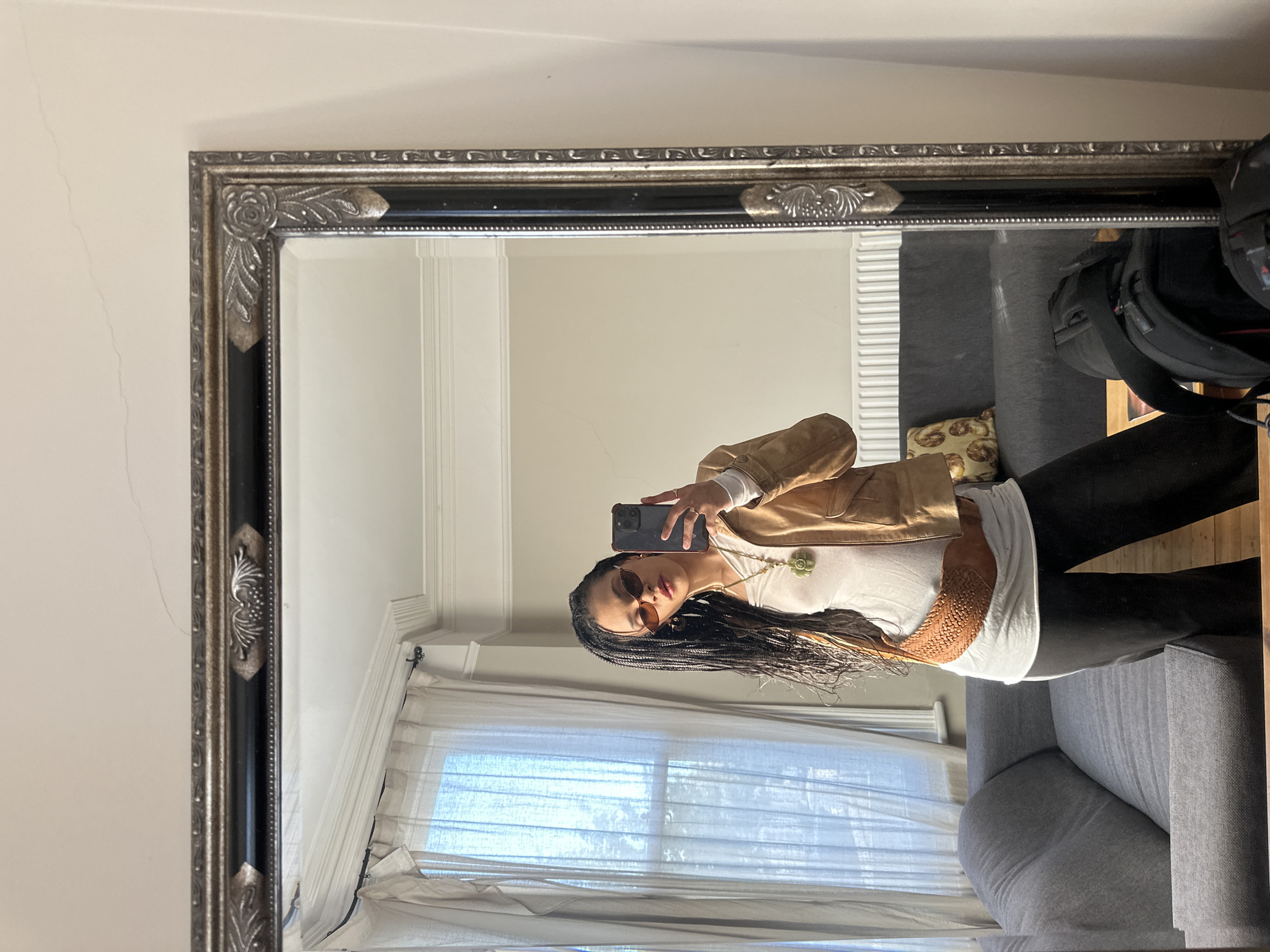 An indoor, well-lit mirror selfie shows a woman posing in what appears to be a living room or bedroom. The image is taken from a slightly high angle, reflecting the woman and a segment of the room she is in.
The central subject is a woman with long, dark braided hair, wearing sunglasses, a white or off-white top, a light brown/tan jacket, and black pants. A green pendant necklace hangs around her neck. She is holding a smartphone with a dark case, angled to capture her reflection in the mirror.
The large rectangular mirror features an ornate frame, dark in color with silver or pewter-toned decorative accents including roses, leaves, and scrolled patterns. Reflected in the mirror behind the woman is a room with a light-colored wall and a window covered by sheer, light-colored curtains, indicating natural light. To the right of the woman, a grey sofa is visible, on which a black bag (possibly a backpack) and a decorative pillow with a light brown, possibly pastry-themed, pattern are placed. Parts of a radiator or wall paneling are also reflected.
The activity is a casual mirror selfie, likely taken during the daytime due to the bright natural light entering through the window. Minor hairline cracks are visible on the wall directly surrounding the mirror. No discernible text is visible in the image. The setting suggests a comfortable, private indoor space in London, United Kingdom.