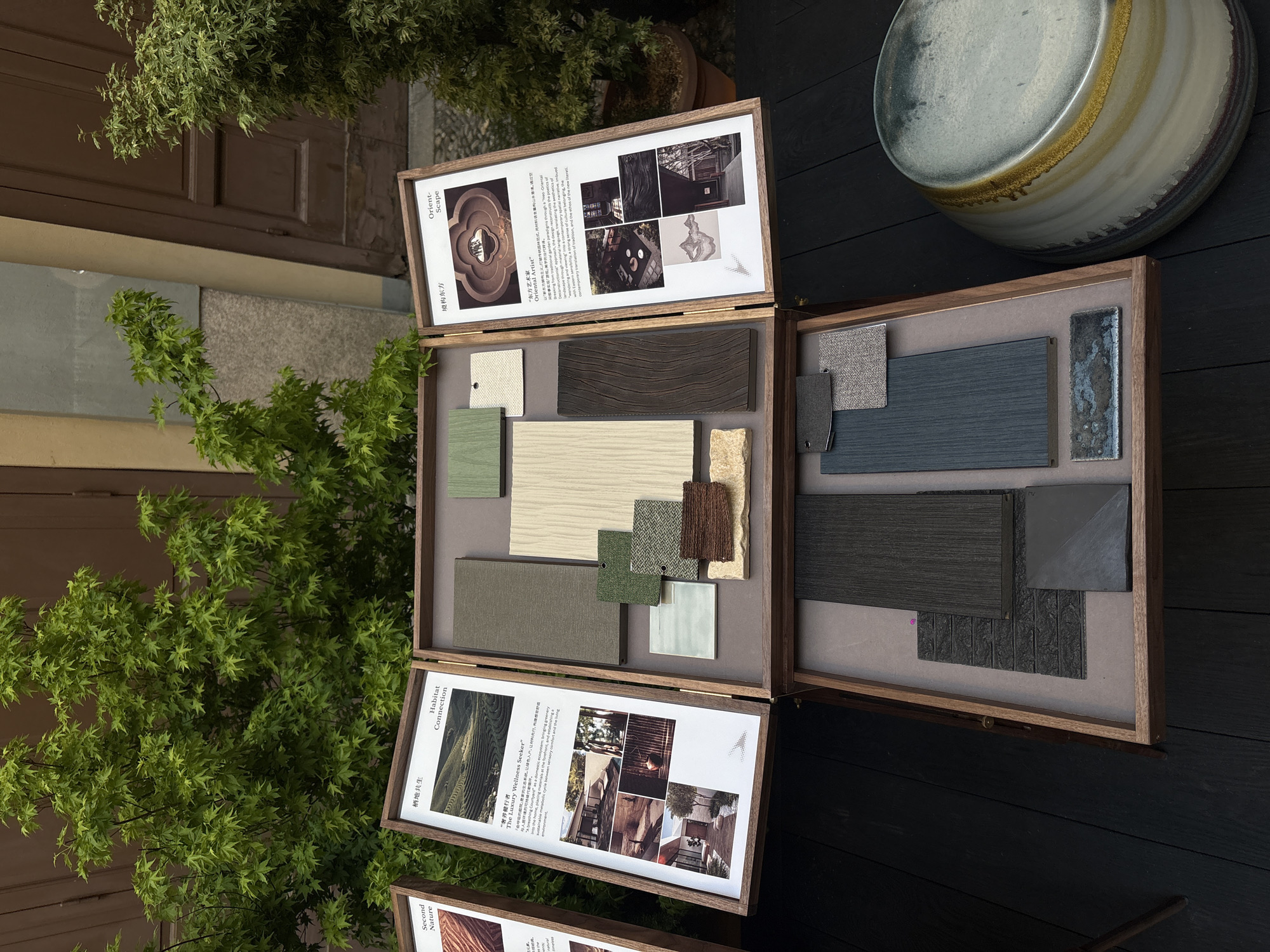 This image captures an outdoor display of design material samples, likely at an exhibition in Milan, Italy. Three open wooden boxes, positioned on a dark wooden surface, showcase various interior and architectural materials.
The boxes are filled with an array of neatly arranged material swatches, including different textures of fabrics, wood veneers, ceramic tiles, stone samples, and metal finishes. The color palette predominantly features earthy tones such as greens, grays, browns, and blues, suggesting a connection to natural elements.
The lids of two of the visible boxes are propped open, revealing printed documents that serve as concept boards or mood boards. The top document is titled "Orient- Scape" (东方艺术家 / "Oriental Artist") and includes text describing a "Neo-Oriental narrative" and "breaking paradigms." It features several small photographs of contemporary interior spaces. The middle box's lid is titled "Habitat Connection" (栖地共生 / "The Luxury Wellness Seeker"), with text mentioning "呼吸的是家的生态系统" (breathing the ecosystem of the home) and "bringing greenery into" living spaces, accompanied by images of nature-integrated architecture. A third box, partially visible at the bottom left, shows the title "Second Nature."
In the upper right corner of the frame, a large, decorative ceramic pot or bowl with iridescent glazing rests on the dark wooden surface. Lush green foliage, possibly from a small tree or bush, frames the scene from the left, while brown wooden doors or panels are visible in the background. The natural lighting indicates a daytime setting. No people are present in the image.