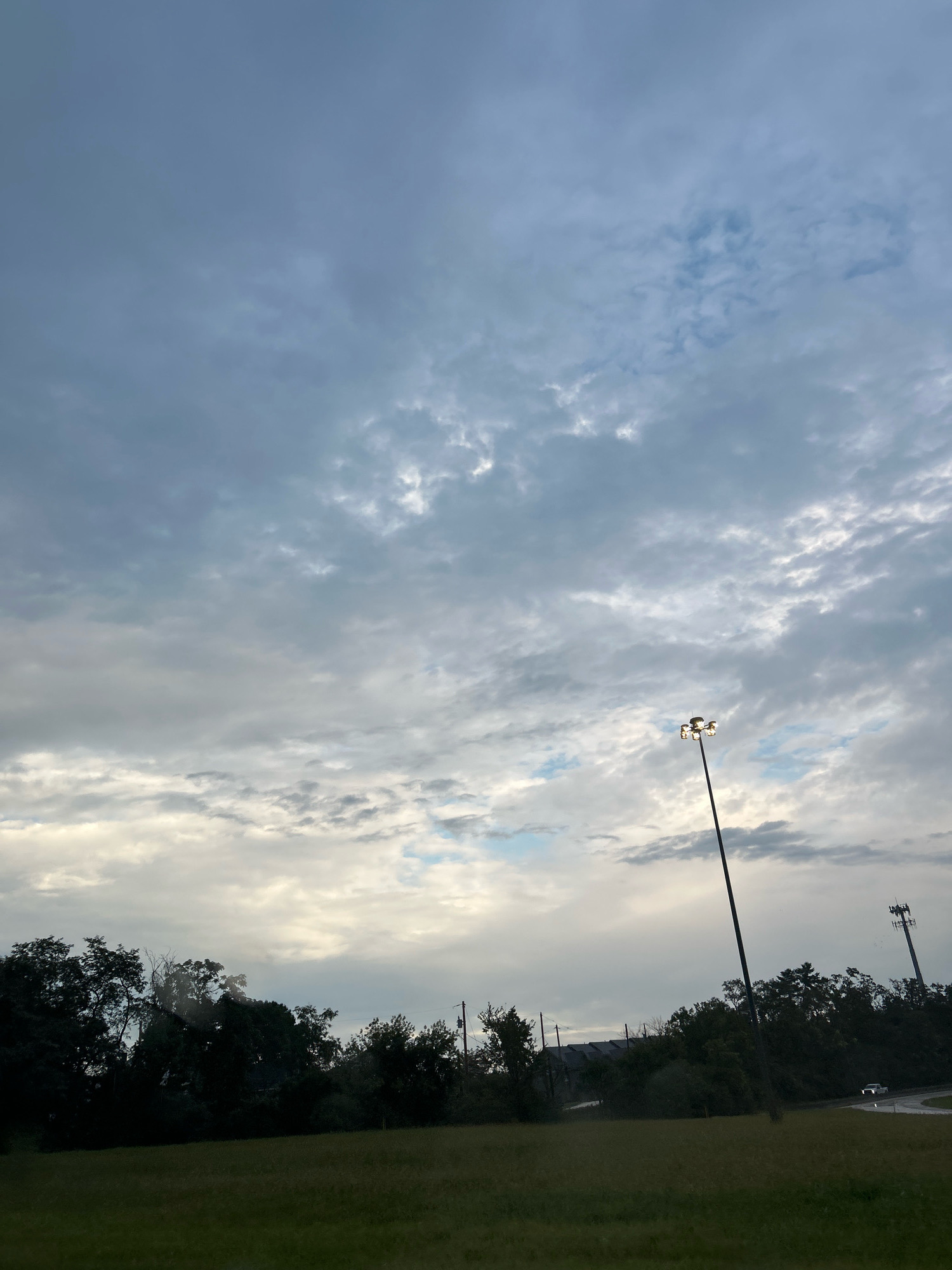 The image depicts an outdoor scene in Robinson Township, United States of America. The sky is overcast with a mix of gray and white clouds, with some patches of blue visible. In the foreground, a grassy field stretches across the lower portion of the frame. A line of dark trees forms a dense silhouette along the horizon. A tall lamppost with multiple lights stands prominently in the middle right of the image, and further to the right, a telecommunications tower is visible. A white vehicle is seen on a road that curves into the distance, partly obscured by trees. The overall lighting suggests it is either early morning or late evening.