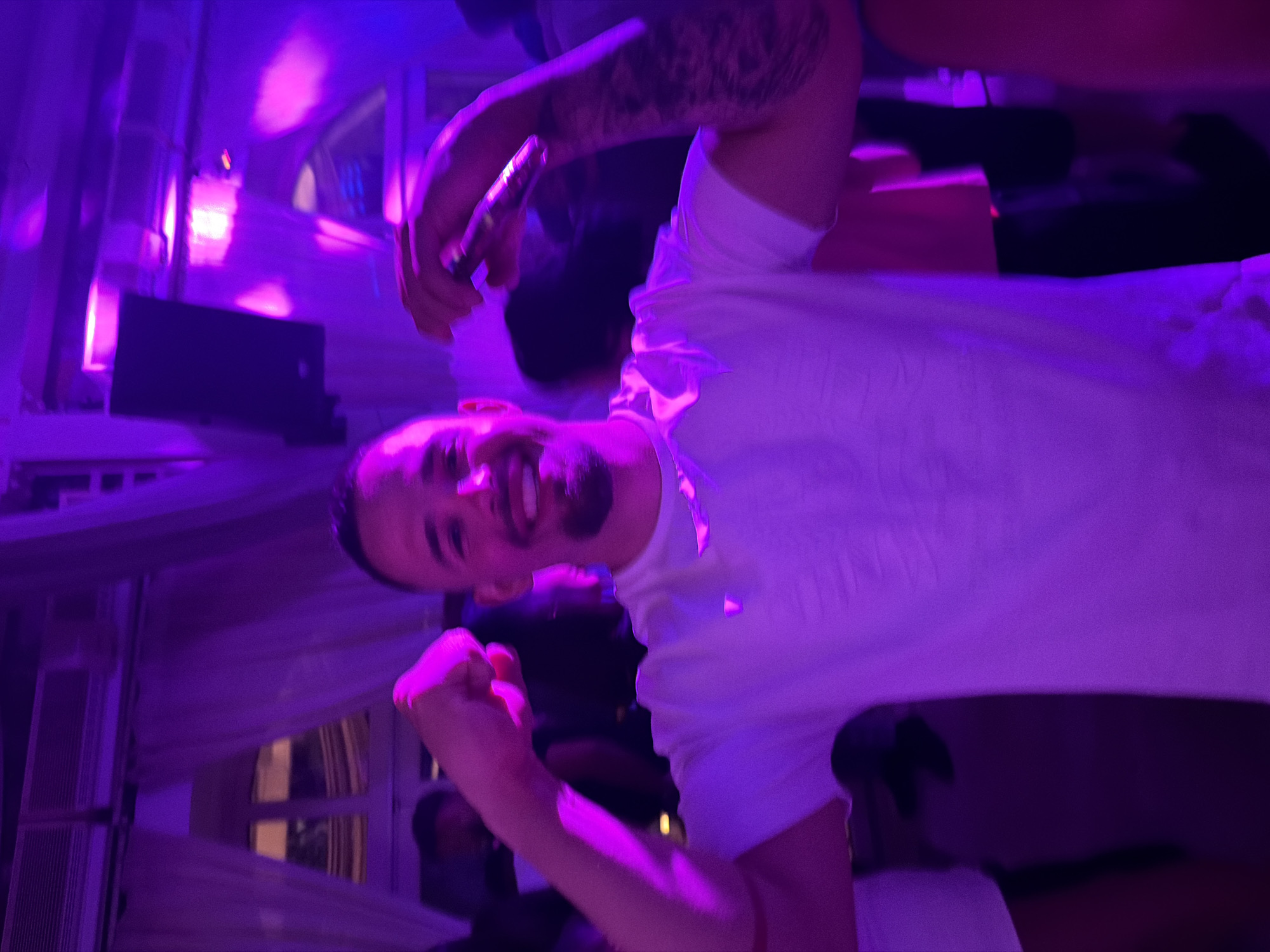 A man in a white t-shirt is holding a microphone and smiling. The image appears to be taken indoors with purple lighting. A window with vertical bars can be seen in the background. Other people are present in the background, but they are out of focus. The location is Pallini Municipal Unit, Greece.