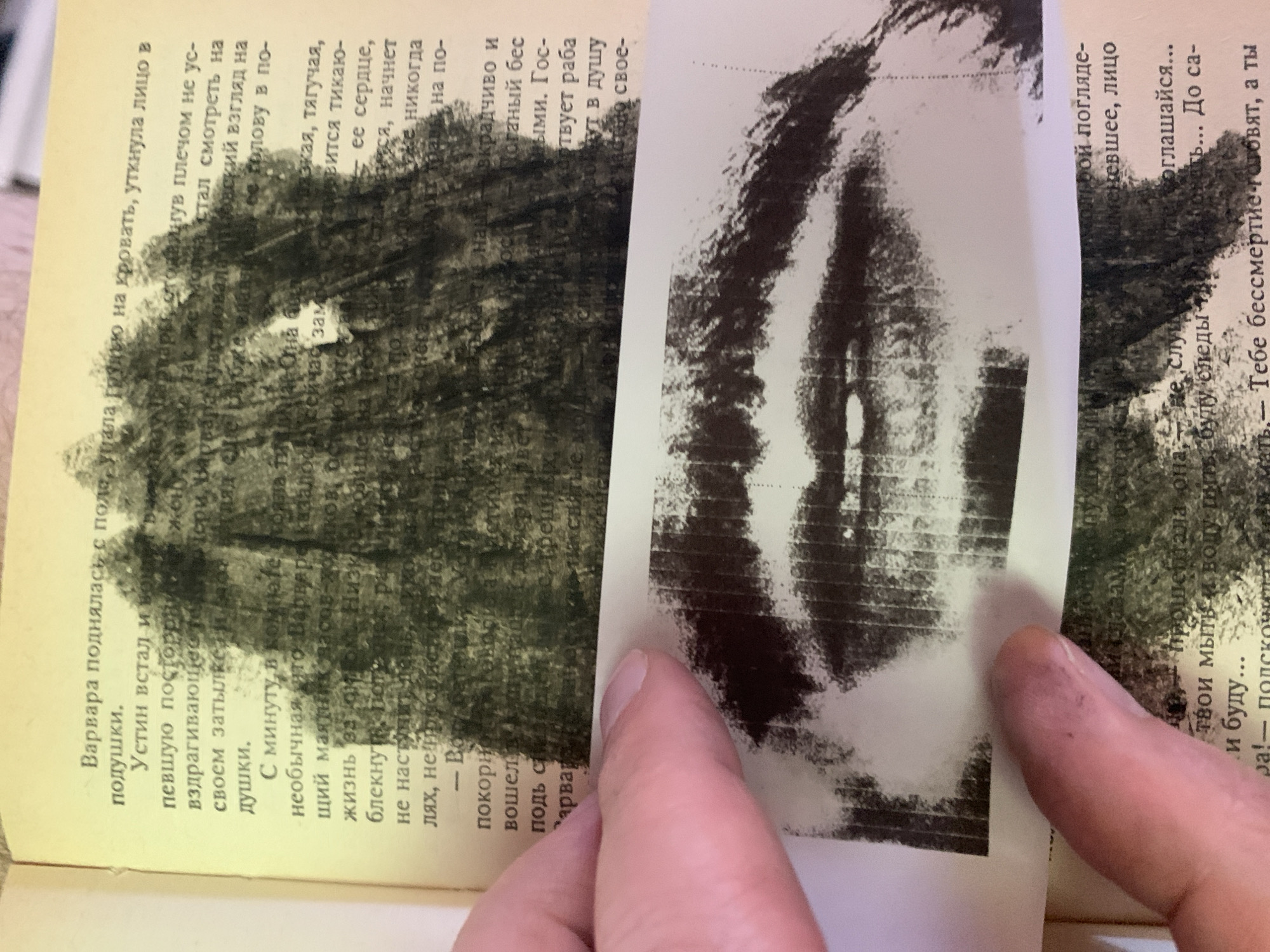 The image is a close-up of an open book with text in Russian. A portion of a black and white drawing or printed image of lips is placed over the text. The book pages are yellowed with age, and a human finger, with a slightly dirty nail, is visible in the lower left corner, holding down the drawing. The text discusses a character named Varvara, who gets up and falls on the bed, and Ustein, who is also mentioned. The drawing of lips is abstract and obscures much of the text, with the lips appearing somewhat smudged. The background appears to be a dark, textured area, possibly part of the drawing or an overlay. There is no visible information in the image that would allow for determining a location in the city of Ganino, Russia.