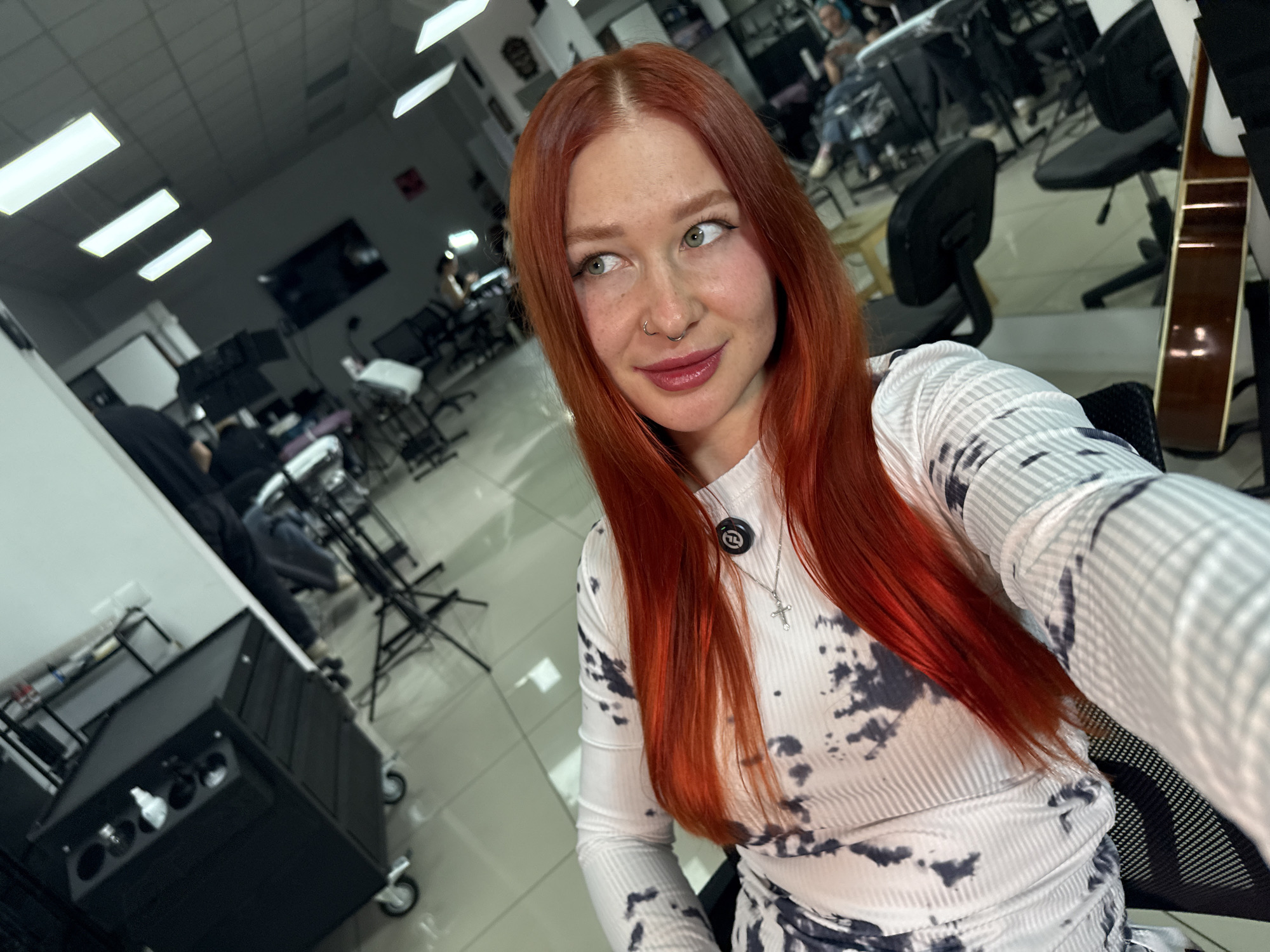 The image is a selfie taken in what appears to be a tattoo parlor or beauty salon in Rostov-on-Don, Russia. The main subject is a woman with long, vibrant red hair, fair skin, and facial piercings. She is wearing a light-colored, patterned long-sleeved top and a necklace with a cross pendant. She is seated, facing the camera with her head slightly turned to the left.
In the background, several other individuals are seated, seemingly receiving services or waiting. There are salon chairs, equipment, and a black rolling cart with various containers. A guitar is leaning against a chair, and overhead lighting is visible. The environment looks to be a commercial space with a functional and slightly industrial aesthetic.