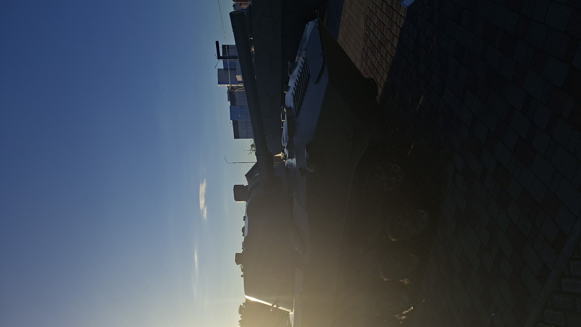 A military tank, possibly a BMP (Boyevaya Mashina Pekhoty - Infantry Fighting Vehicle), is parked on a cobblestone surface in front of buildings under a clear blue sky. The sun is low on the horizon, casting a strong backlight and creating silhouettes. The tank is dark, with some of its features visible, including its tracks, wheels, and turret with a cannon. In the background, several modern, multi-story buildings with windows are visible, along with some streetlights. The scene suggests an outdoor display or storage area in a city. The location is identified as Smaliavichy, Belarus.