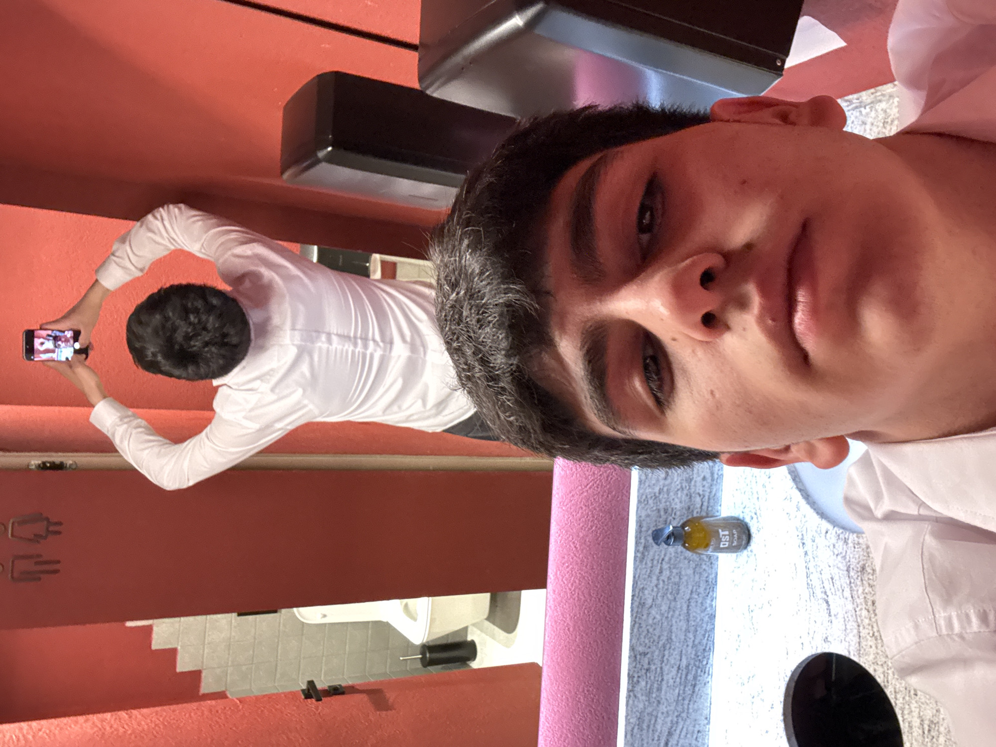 A man takes a selfie in a restroom, possibly in Yerevan, Armenia. He has dark hair and is wearing a white collared shirt. In the background, a man is taking a picture of the restroom mirror. The restroom has red walls, white fixtures, gray countertops, and a soap dispenser. A black step stool sits near the countertop. The floor appears to be tiled. Restroom signage is visible on the wall.