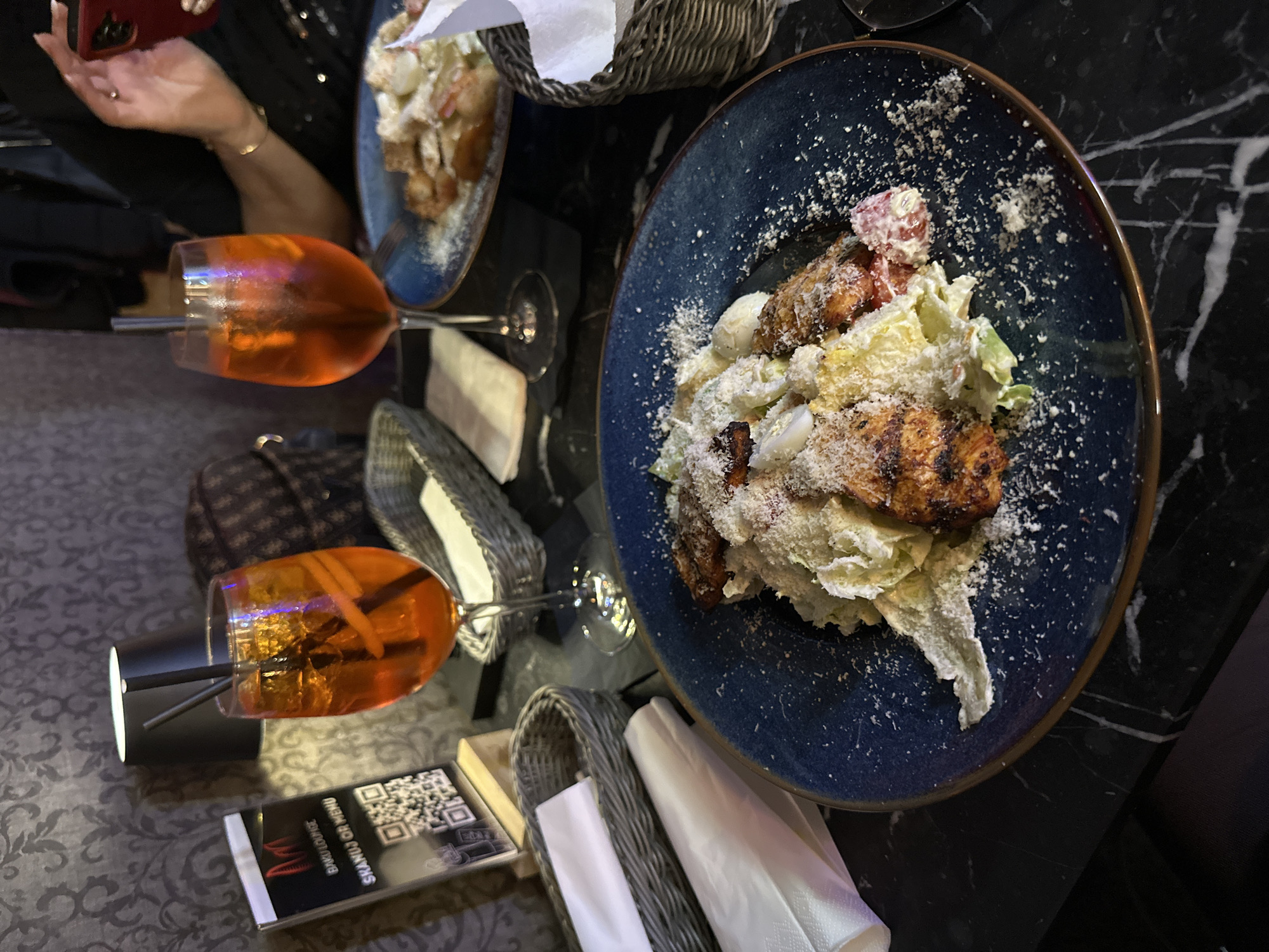 The image depicts a restaurant table setting, likely in Wrocław, Poland, given the menu card visible on the table which states "Baku Lounge, Shamuaz or Menu". Two Aperol Spritz cocktails are present in stemmed glasses, along with two plates of Caesar salad, one closer to the viewer and one in the background. Cutlery and napkins are arranged in wicker baskets. The table has a patterned surface with a visible marble section. One person’s hand holding a smartphone is visible in the upper left.