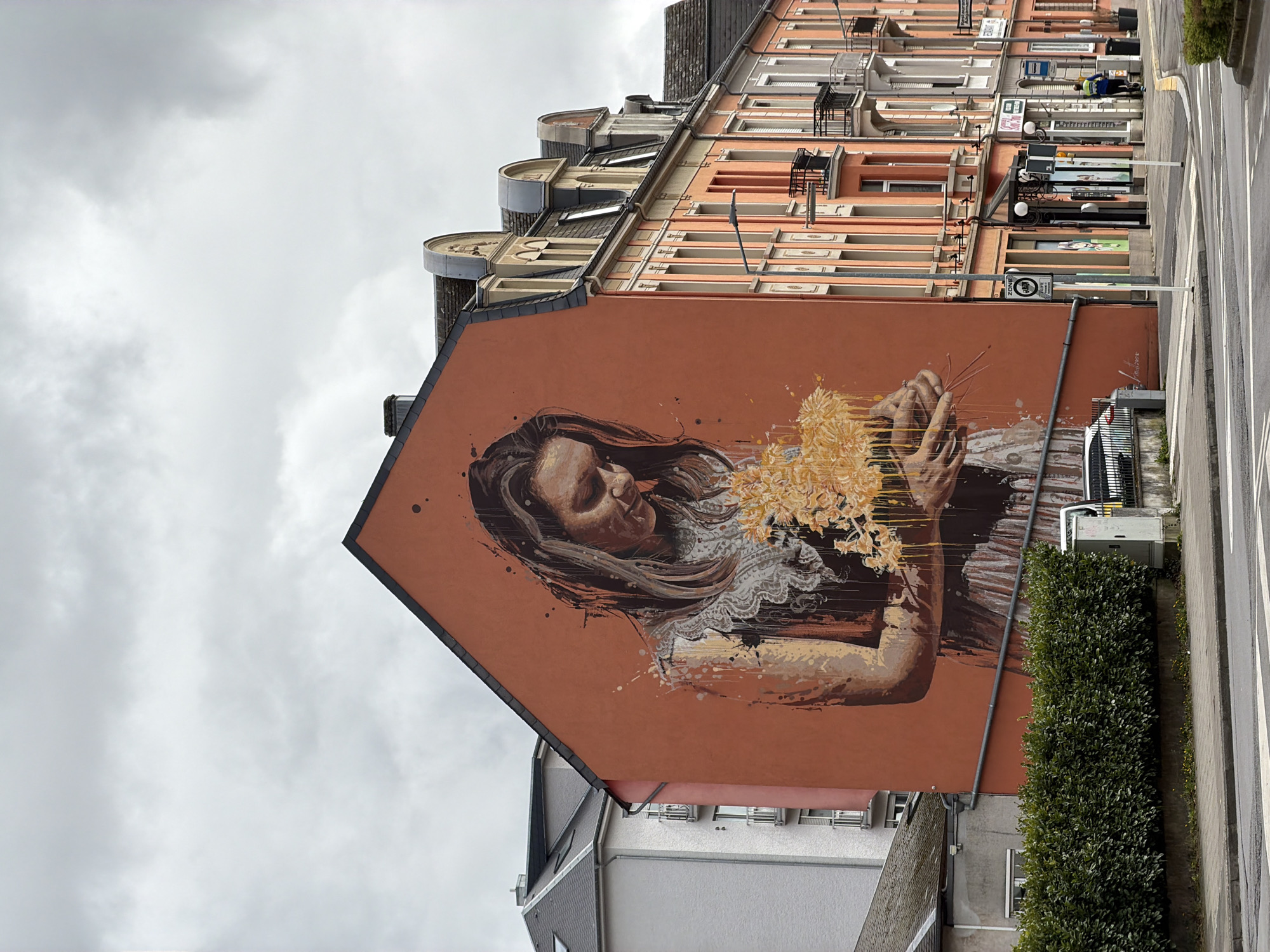 A mural of a woman holding yellow flowers is painted on the side of a building in Esch-sur-Alzette, Luxembourg. The woman has long brown hair and is wearing a lace-trimmed top. The building is located on a street with other buildings and greenery.