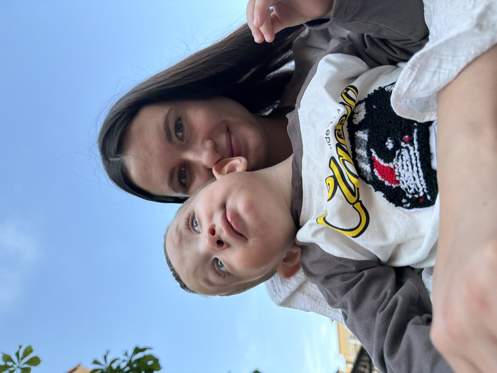 This is a close-up, low-angle shot of a young woman holding a baby. They are outdoors against a clear blue sky. The woman, with long dark hair and fair skin, is looking down at the baby and smiling. Her eyes are wide and her lips are parted in a happy expression. The baby, who appears to be a toddler, is looking up at the woman with a playful expression, his tongue sticking out slightly. He has light skin and light blue eyes. The baby is wearing a white t-shirt with a black and white cartoon-like graphic of a face with red accents. The woman is wearing a gray top.
The scene suggests a candid moment of interaction and affection between the mother and child. The lighting indicates it is daytime, likely sunny. The presence of greenery at the bottom of the frame suggests an outdoor setting, possibly a park or a backyard. There is no visible text in the image.
