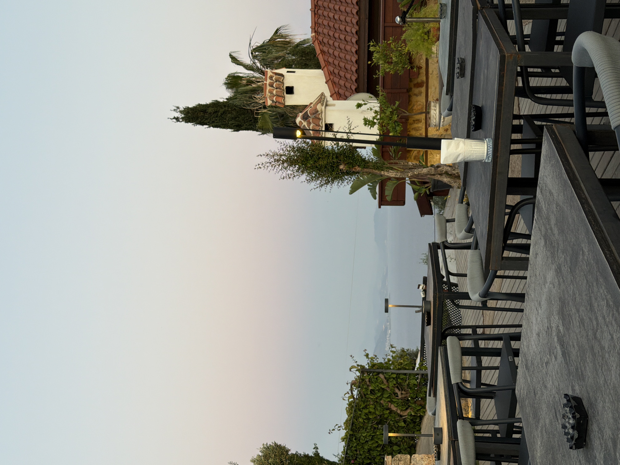 The image shows an outdoor dining area in Alanya, Turkey, likely part of a restaurant or cafe. The area features several dark-colored tables and chairs arranged on a wooden deck. A building with a red tile roof and white walls is visible in the background, along with various trees and plants. The sky is light and appears to be near dusk or dawn. A body of water, possibly the Mediterranean Sea, can be seen in the distance. Each table has ashtrays. There are no visible people in the scene.
