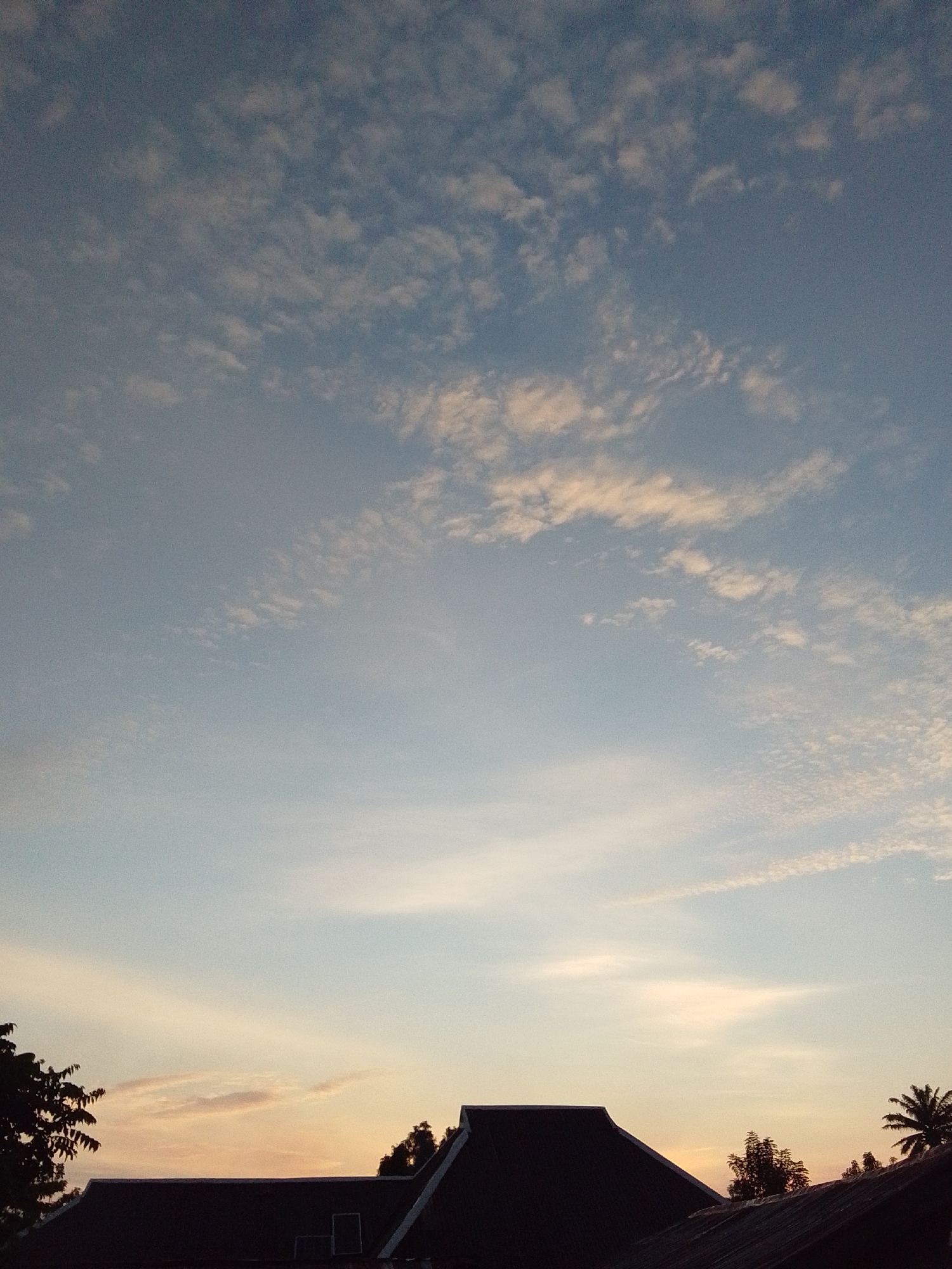 The image captures a twilight sky over Serti, Nigeria. Silhouetted rooftops and trees dominate the lower portion of the frame, indicating an urban or semi-urban setting. The sky is a gradient of soft blues and pale oranges, with scattered, wispy clouds catching the fading sunlight. The overall atmosphere is serene and peaceful, characteristic of dawn or dusk in a tropical region. There are no people or specific actions depicted.