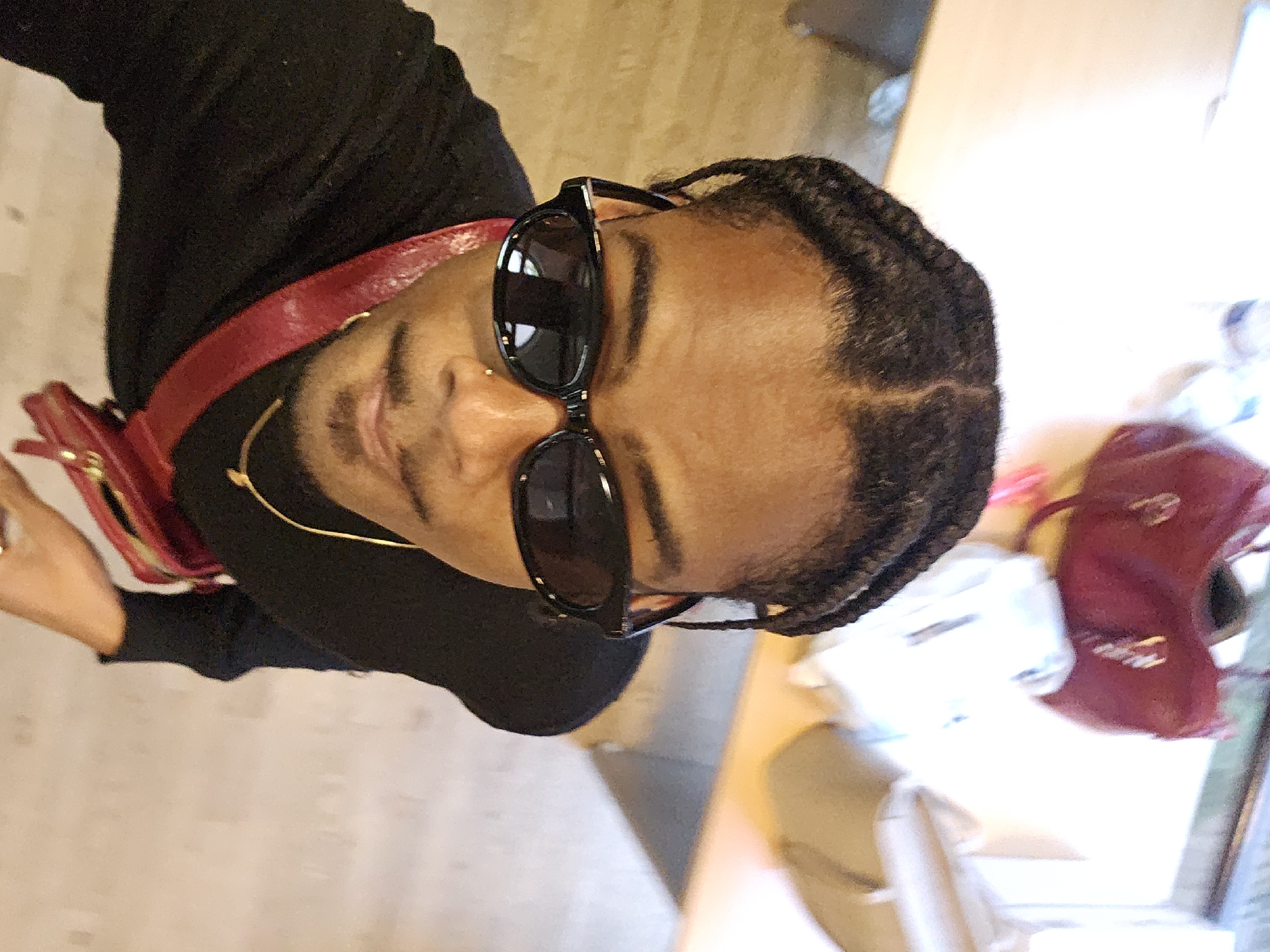 A person with dark hair braided close to the scalp is wearing black sunglasses and a black top. A red purse is slung across their shoulder. They appear to be indoors, possibly in a cafe or restaurant, given the table and items in the background. The exact location in Paris is not discernible from the image.