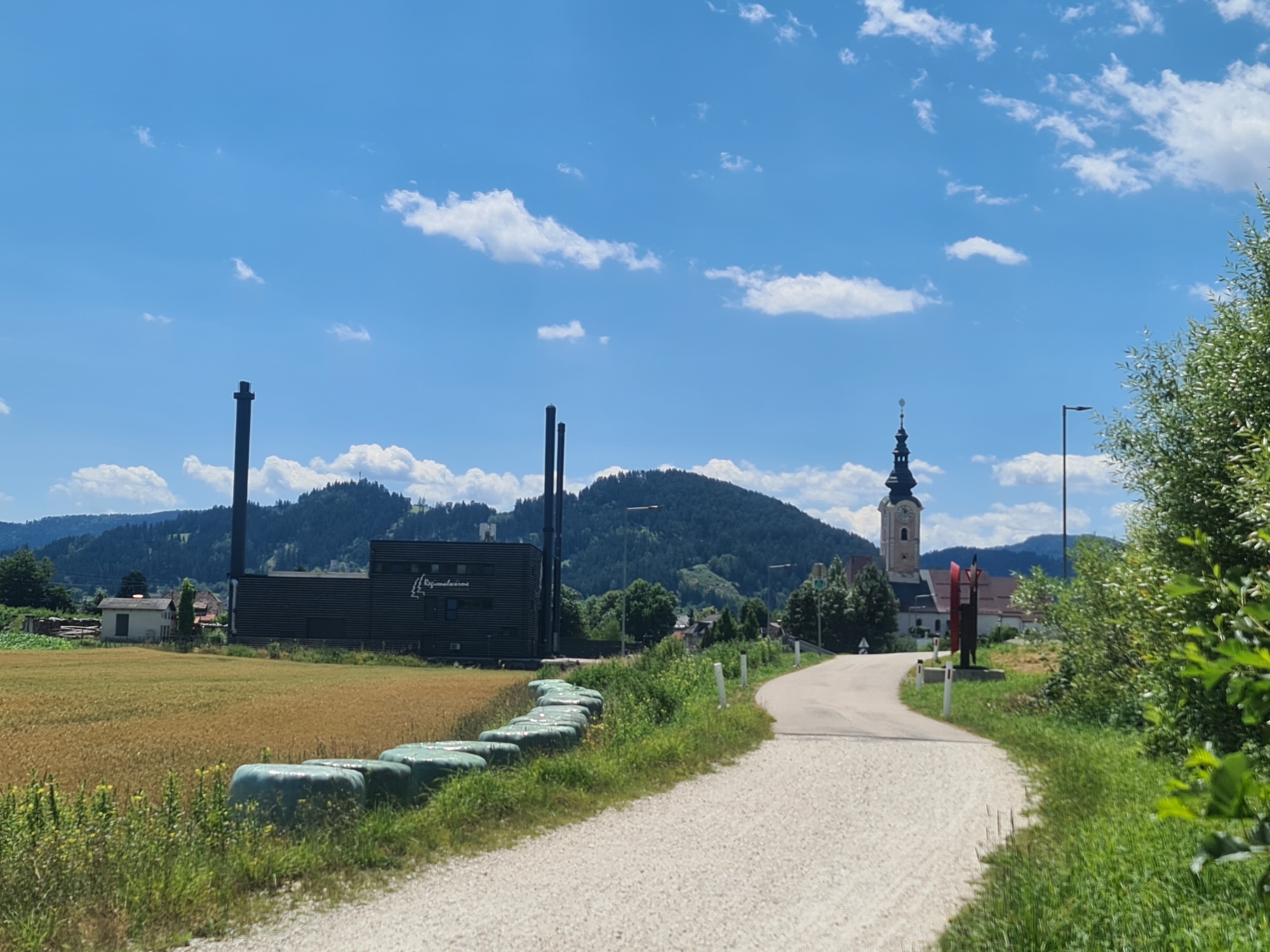The image depicts a rural scene on a sunny day, likely in or near Feldkirchen in Kärnten, Austria. In the foreground, a gravel road curves to the right, bordered by green grass and a row of round bales of hay wrapped in green plastic. Beyond the bales, a field of golden crops, possibly wheat, stretches to the left.
Dominating the mid-ground is a large, dark-colored industrial building with several tall smokestacks. The building has "Regionalwärme" written on its facade. Behind the industrial building, a picturesque green hill rises, dotted with trees and some smaller structures.
To the right of the industrial building and closer to the road, a church steeple with a clock is visible, indicating a settlement. The church is adjacent to other buildings. A tall, dark metal pole with a light fixture stands on the right side of the road. In the far background, more rolling hills covered in dense forest can be seen under a bright blue sky with scattered white clouds. The overall impression is one of a tranquil, semi-industrialized rural landscape.