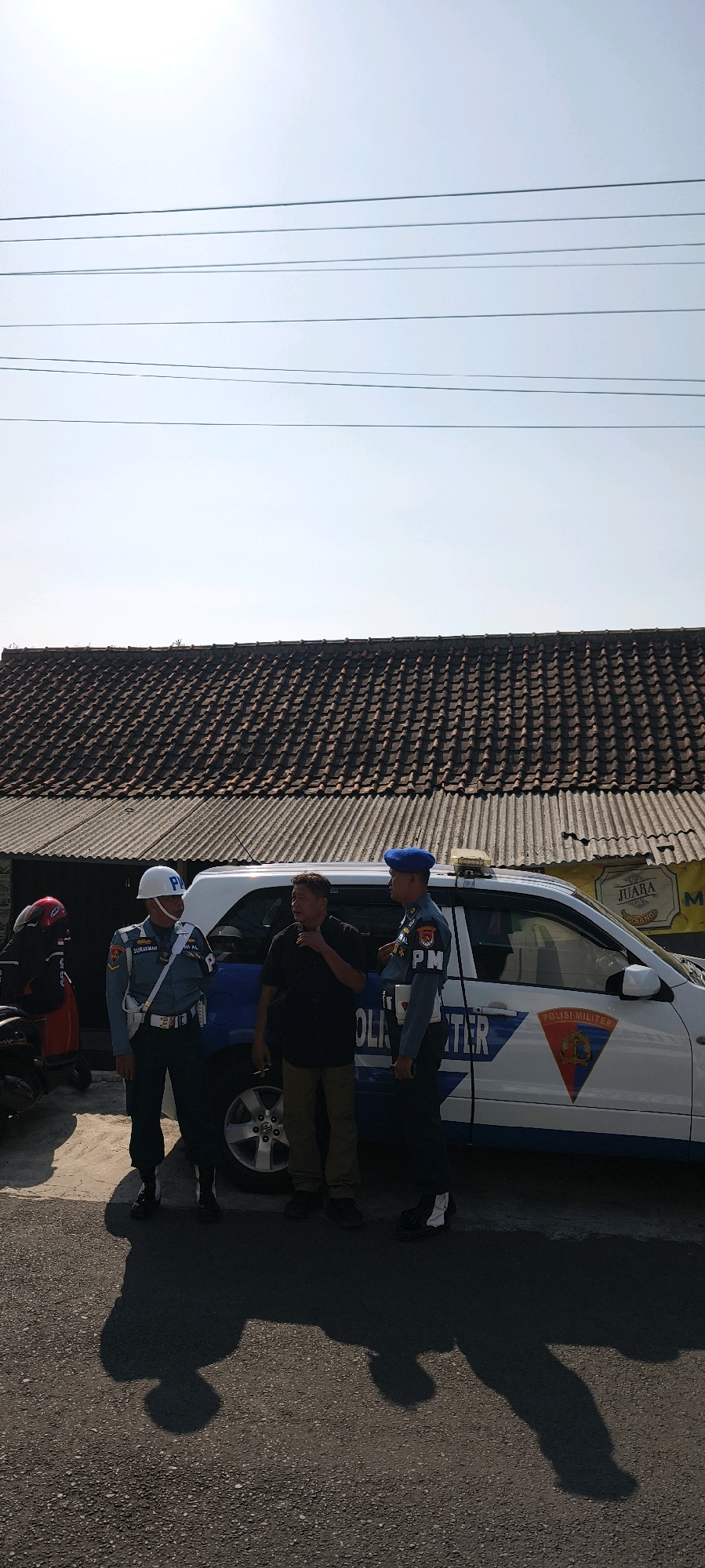 In Sadamantra, Indonesia, on a sunny day with a clear sky and visible overhead power lines, a scene unfolds outside a building with a tiled roof. Three men are present. Two are in uniform, appearing to be military police, distinguished by their blue berets and "PM" insignia on their uniforms. One uniformed man on the left wears a white helmet with "PM" visible. The third man, dressed in a dark shirt and light brown pants, appears to be a civilian and is smoking a cigarette. They are standing near a white SUV with blue markings that include the text "POLISI MILITER" and a shield emblem. The SUV is parked on an asphalt road, and shadows are cast on the ground. A red scooter is partially visible to the left, and what appears to be part of another vehicle behind the uniformed man on the left. The overall impression is of a law enforcement or official interaction taking place outdoors.