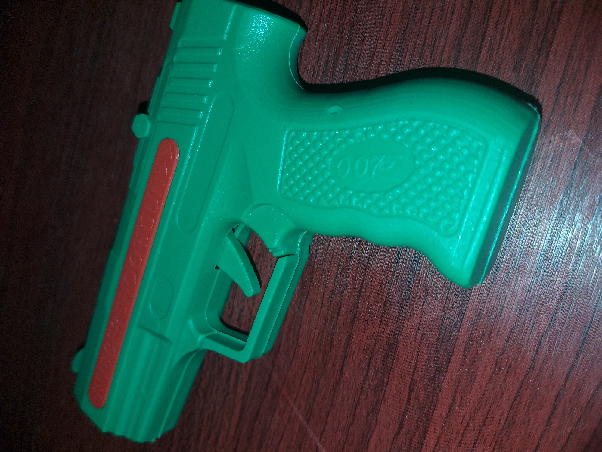 A green toy pistol with a red insert is placed on a wooden surface. The pistol has the inscription "007" on its grip. The wooden surface is dark red and has a visible wood grain. The image is a close-up shot, and the background is dark. There are no people or any other objects visible in the scene. The location is not specified in the image.