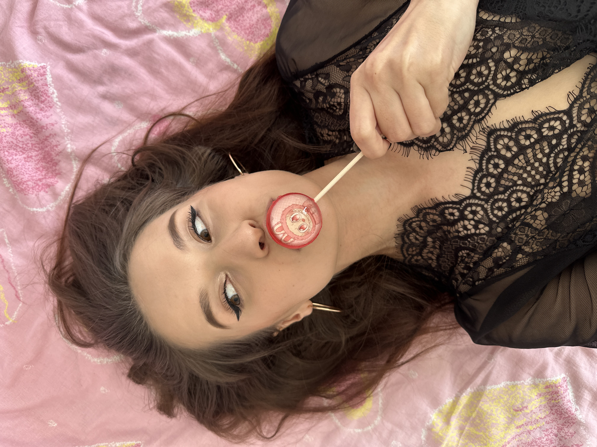 A young woman with long, dark hair lies on a pink bedsheet adorned with light pink, yellow, and white abstract floral patterns. She is looking directly at the camera with a neutral expression. Her eye makeup is prominent, featuring winged eyeliner and defined eyebrows. She wears gold-colored hoop earrings. The woman is dressed in a black lace and sheer top, with details of the lace visible on her chest and arm. Her left hand is near her neck, holding a lollipop stick. A red, circular lollipop, depicting a cartoon-like pizza design, is held between her lips. The scene is well-lit, suggesting an indoor setting, possibly in Yekaterinburg, Russia. There is no visible text in the image.