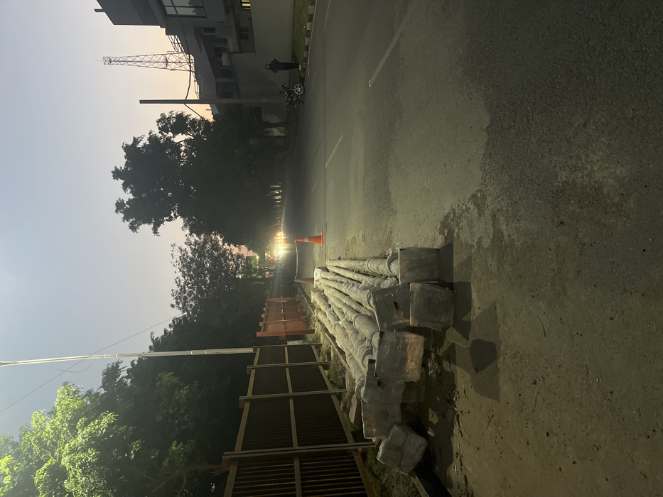 A street in Sahibzada Ajit Singh Nagar, India, at the intersection of two roads, with a row of trees on either side. A metal fence runs along the left side of the street, with several large metal pipes stacked against the fence. In the middle of the street, a traffic cone stands in the center lane, with a chain running across the street, extending from the fence to the opposite side. There are two cars in the distance at the end of the street, as well as one person walking on the opposite side of the street, facing the camera. The sky is twilight, and the ground is covered in dirt and gravel. The street is lit by a street lamp on the left side of the frame. The scene appears to be part of a construction project.
