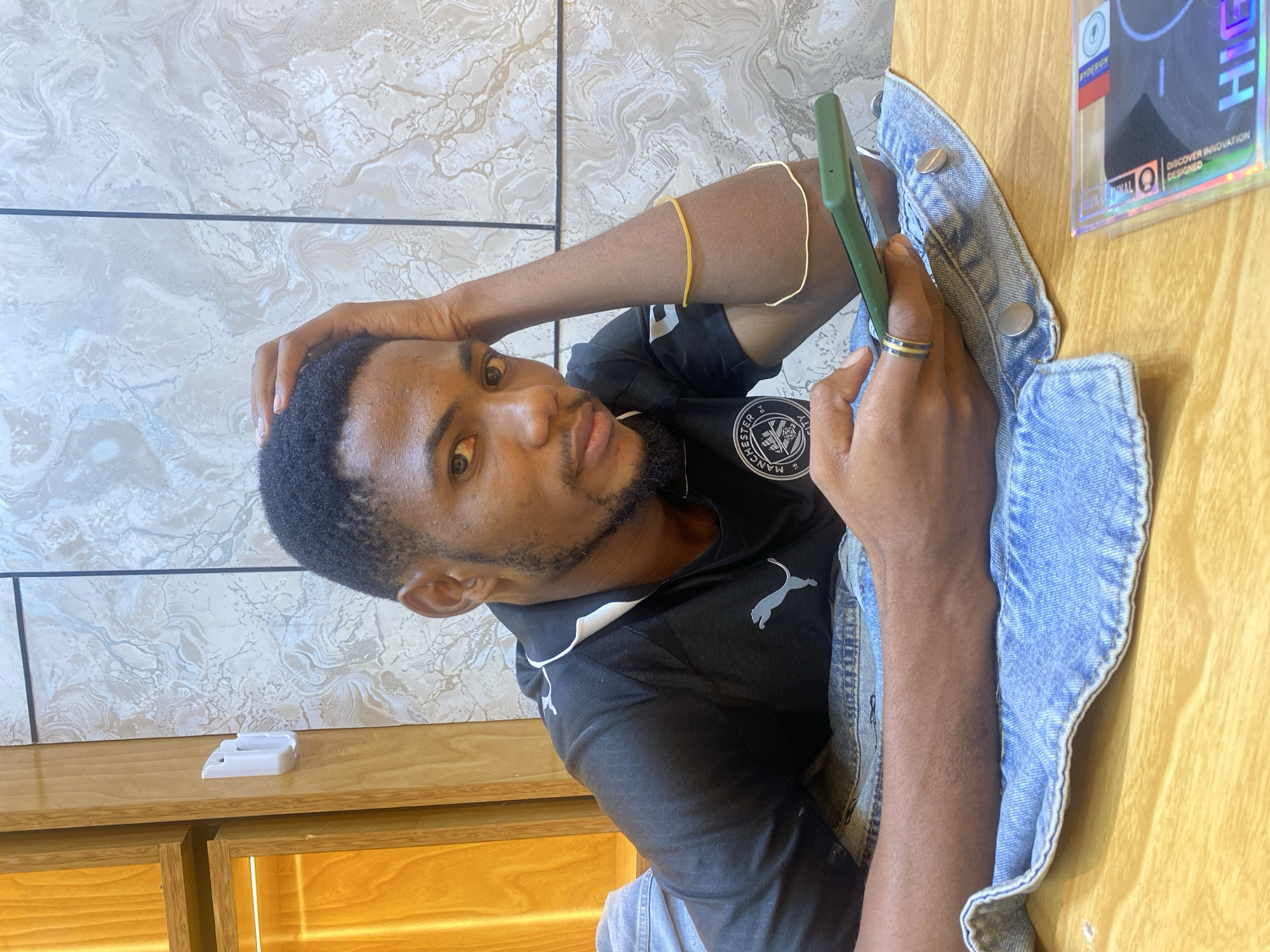 A man is seated at a table indoors. He has short, dark hair and a beard and is wearing a black t-shirt with a Puma logo and a Manchester City crest on the front. He is holding a green cell phone in his right hand. He has a gold ring on his finger and a gold bracelet on his left arm. A light blue denim jacket is draped on the table. The table is wooden. A white object and a package labeled "High By Design" are on the table. The background consists of a wall with a textured pattern. The location is likely a restaurant or cafe in Dar es-Salaam, Tanzania, but this cannot be determined from the image alone.